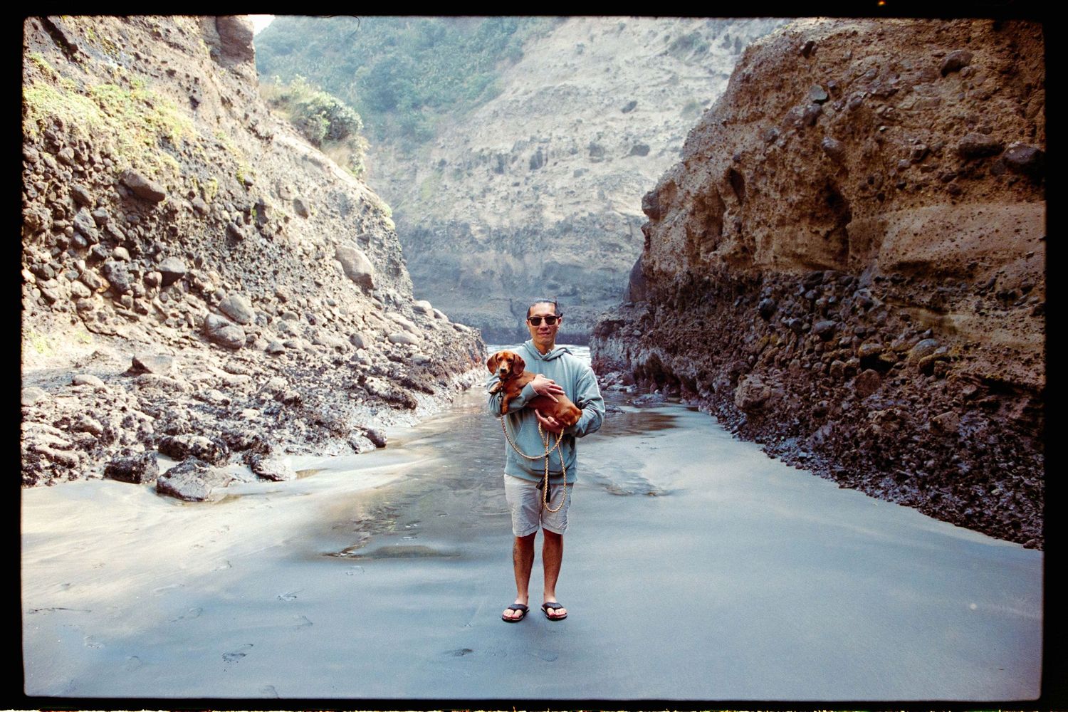 A man in shorts and sandals stands on a wet sandy riverbed holding a small brown dog between steep rocky canyon walls.