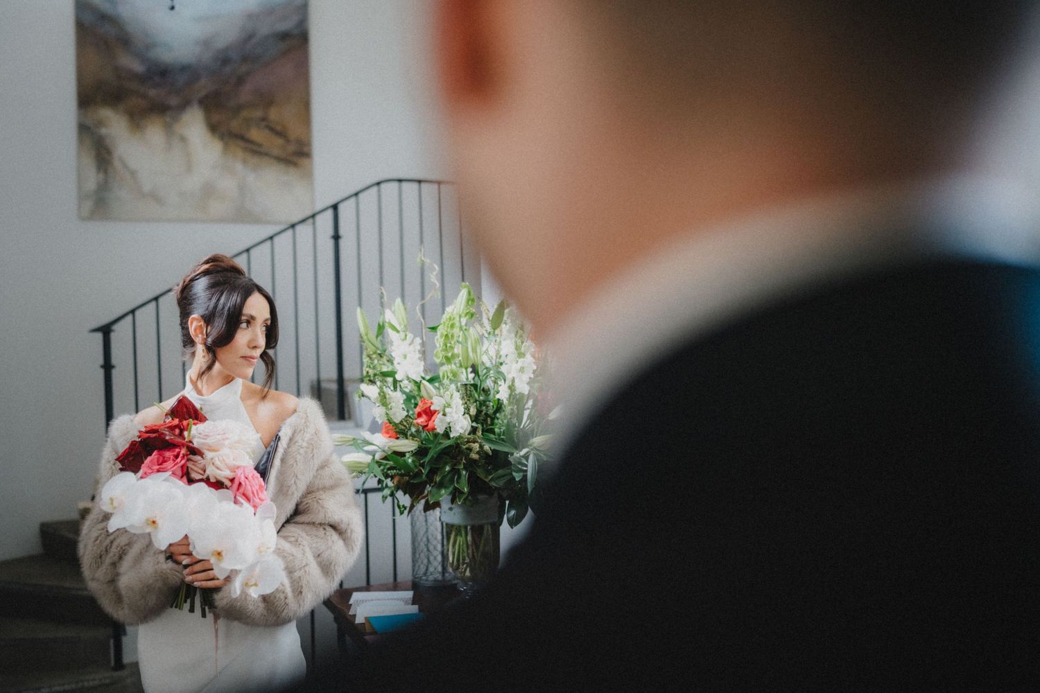 A bride in a cozy shawl holds a red and white bouquet while a blurred groom stands in the foreground.