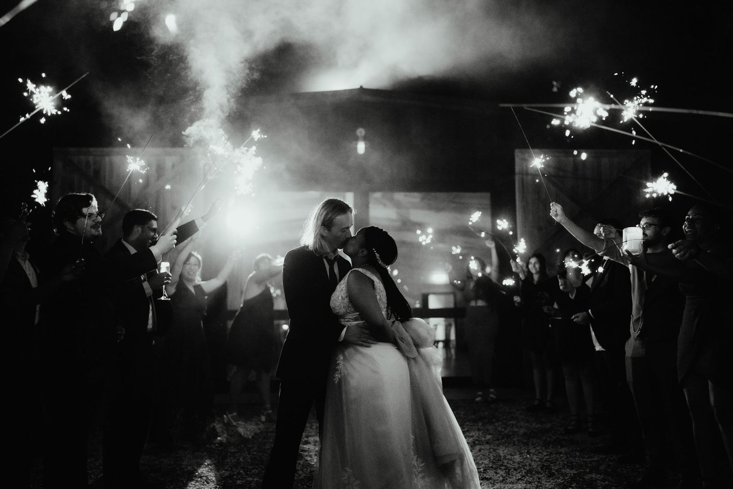 Black and white photo of a bride and groom kissing as wedding guests hold sparklers around them at night.