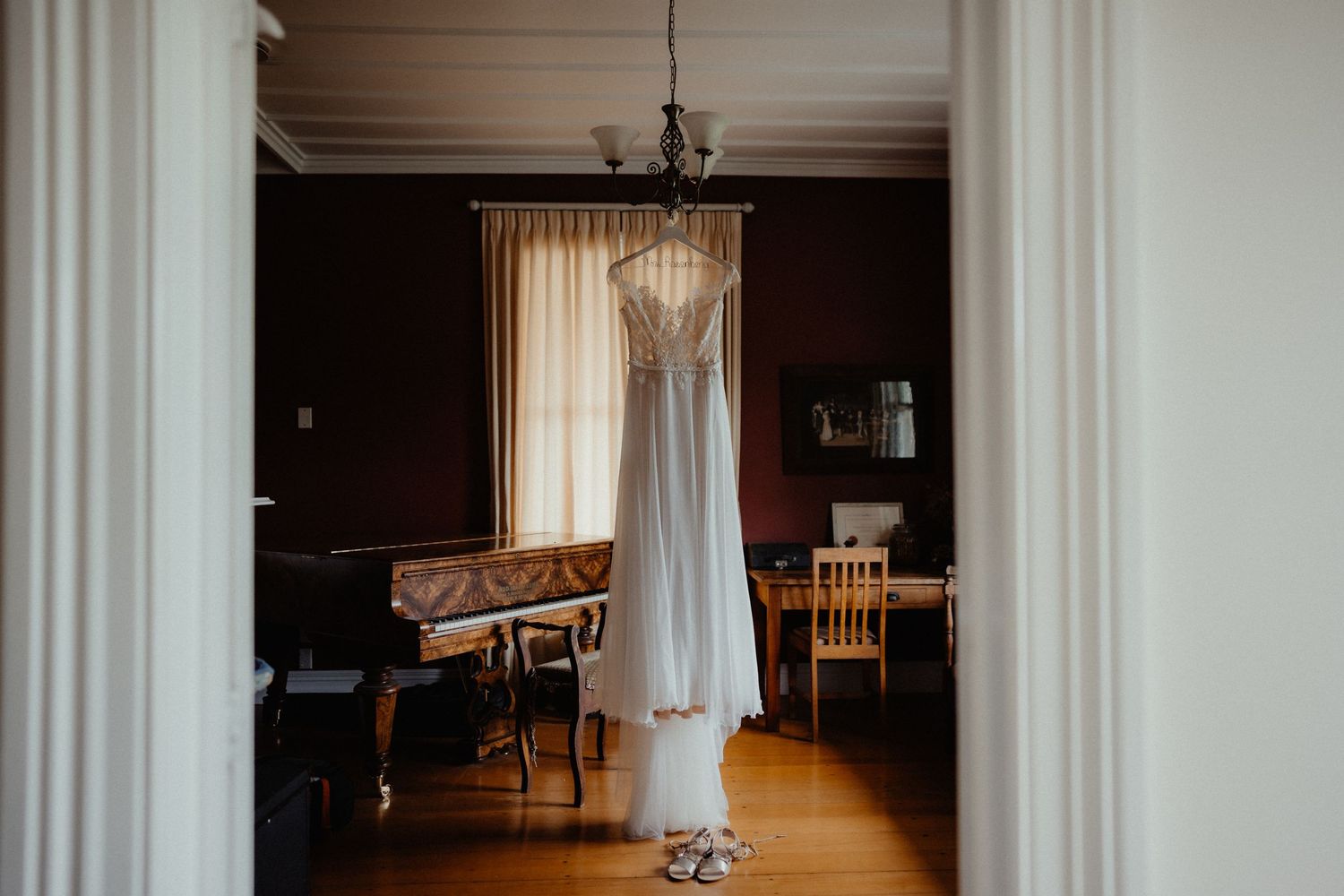 Lace wedding dress hangs from a chandelier in a elegant room with a piano, with the bride's shoes on the floor below.