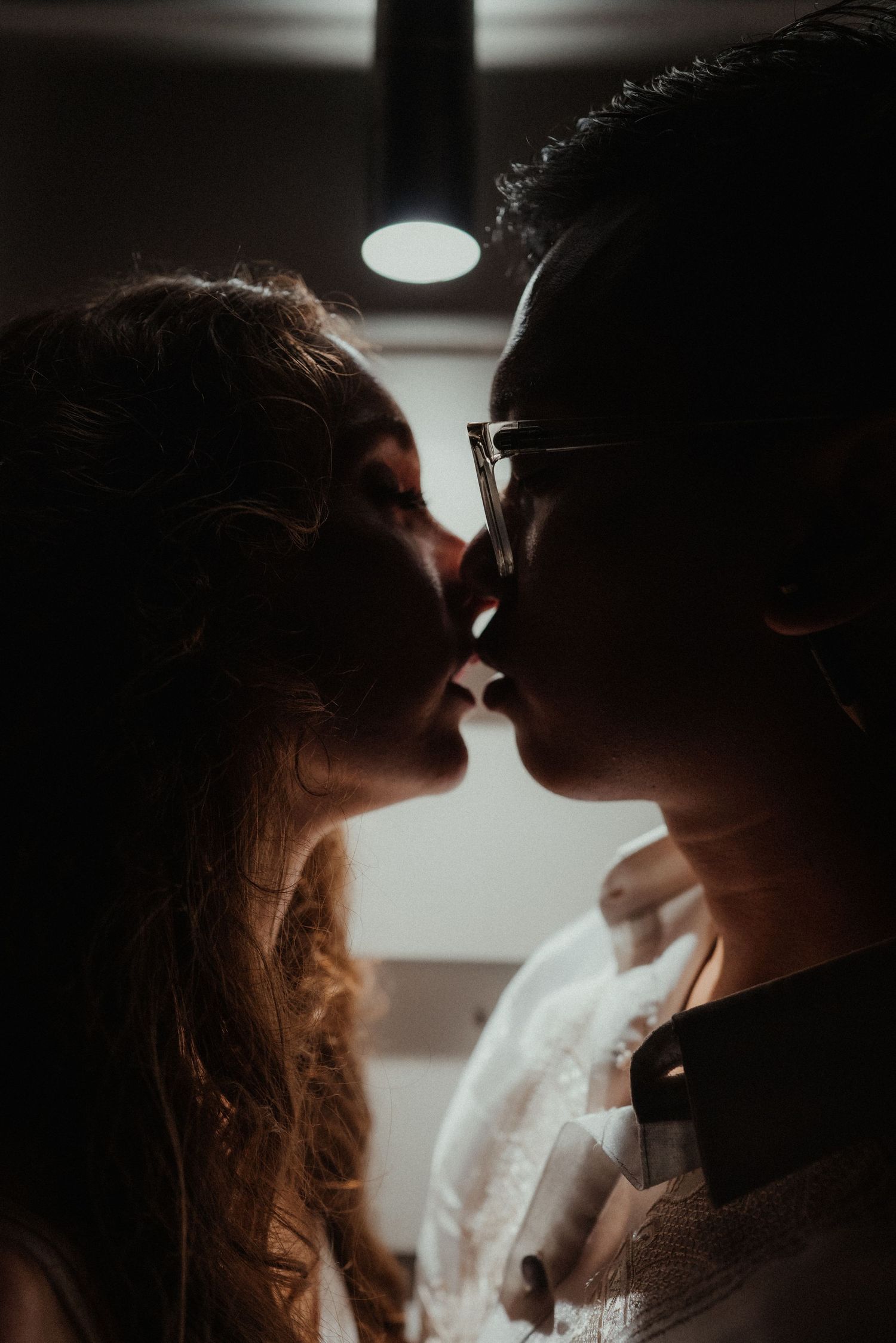 Dramatic silhouette of a couple sharing a kiss in warm, low light, with the groom wearing glasses and the bride's curls visible.