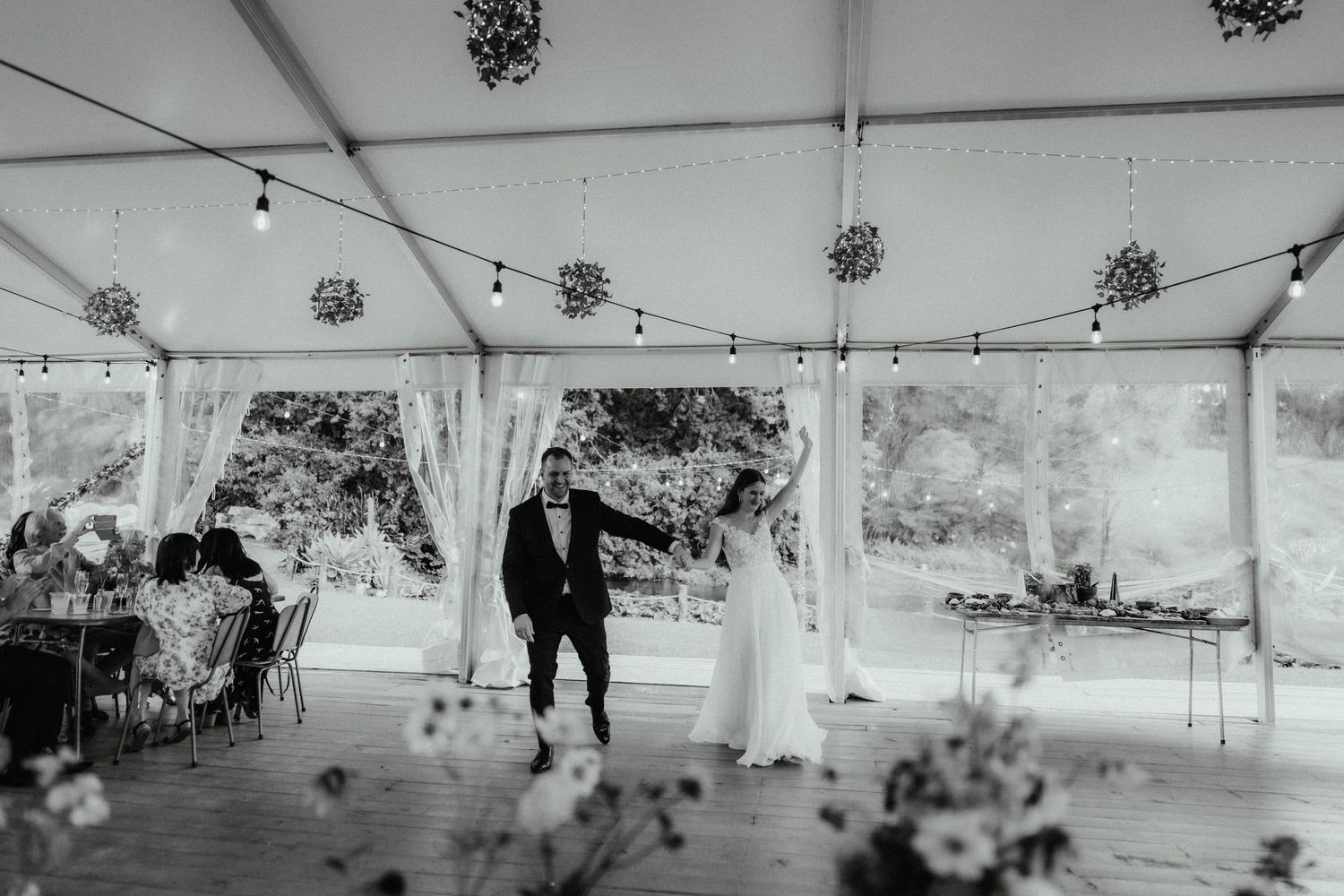 Bride and groom share a joyful first dance in a marquee with string lights while guests watch from tables.