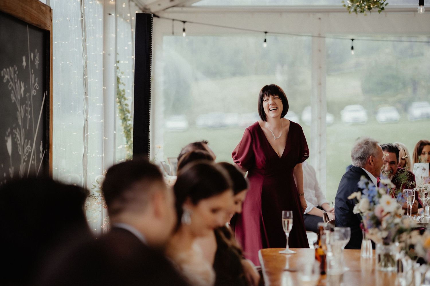 Smiling woman in a burgundy dress gives a wedding speech beside a chalkboard while guests listen at a long reception table.