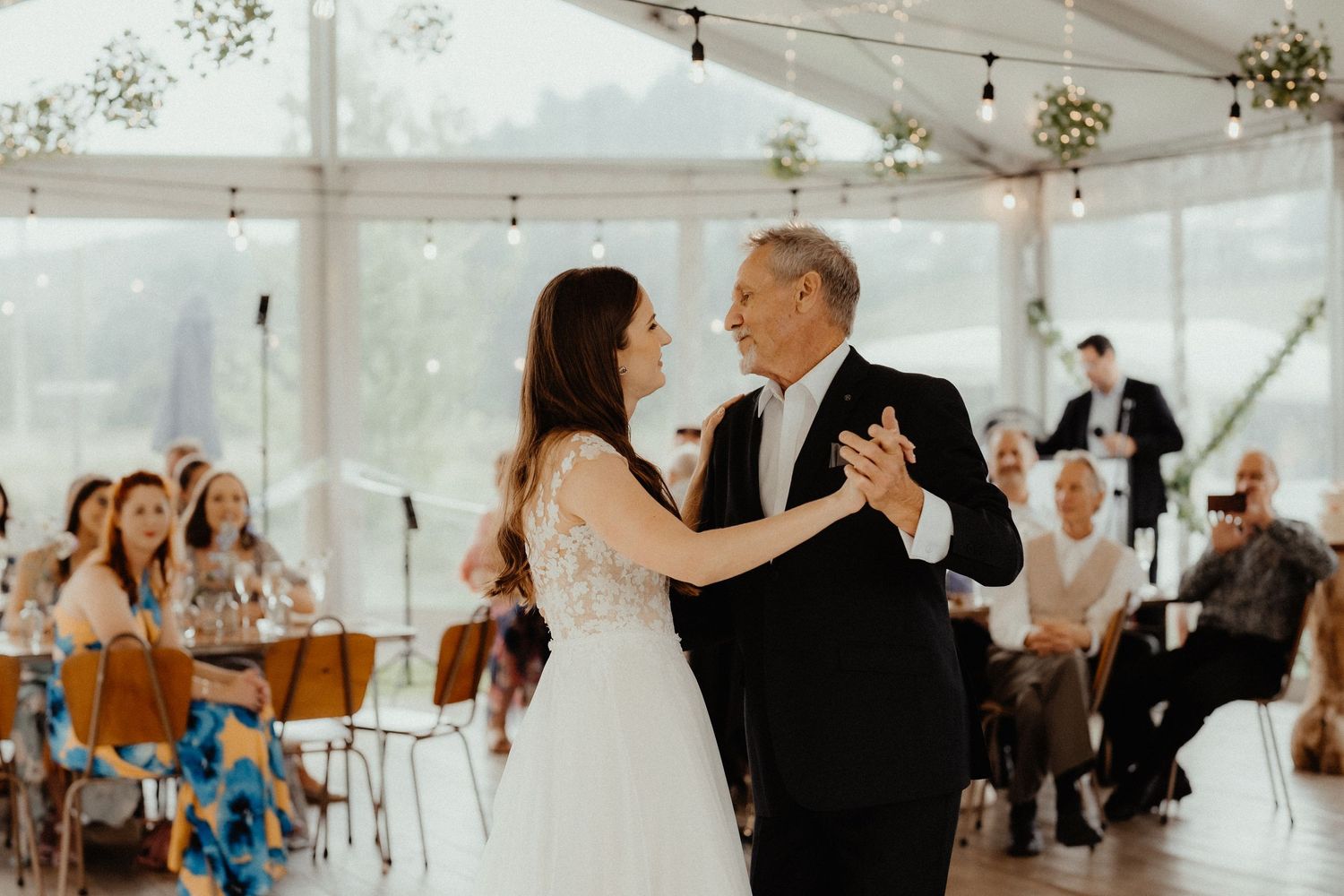 Bride dances with an older man in a black suit at a wedding reception while guests watch from their seats.