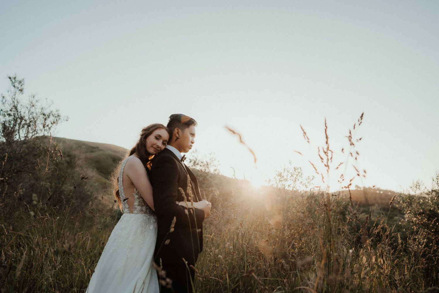Bride in a lace wedding gown embraces the groom from behind in a golden-lit grassy field at sunset.