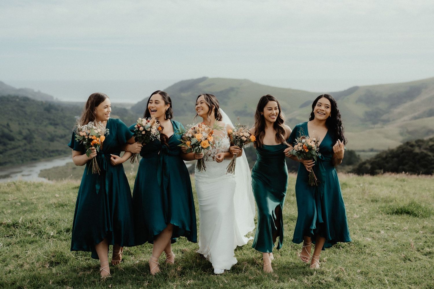 Bride walks laughing with four bridesmaids in emerald green dresses holding bouquets across a grassy hilltop.