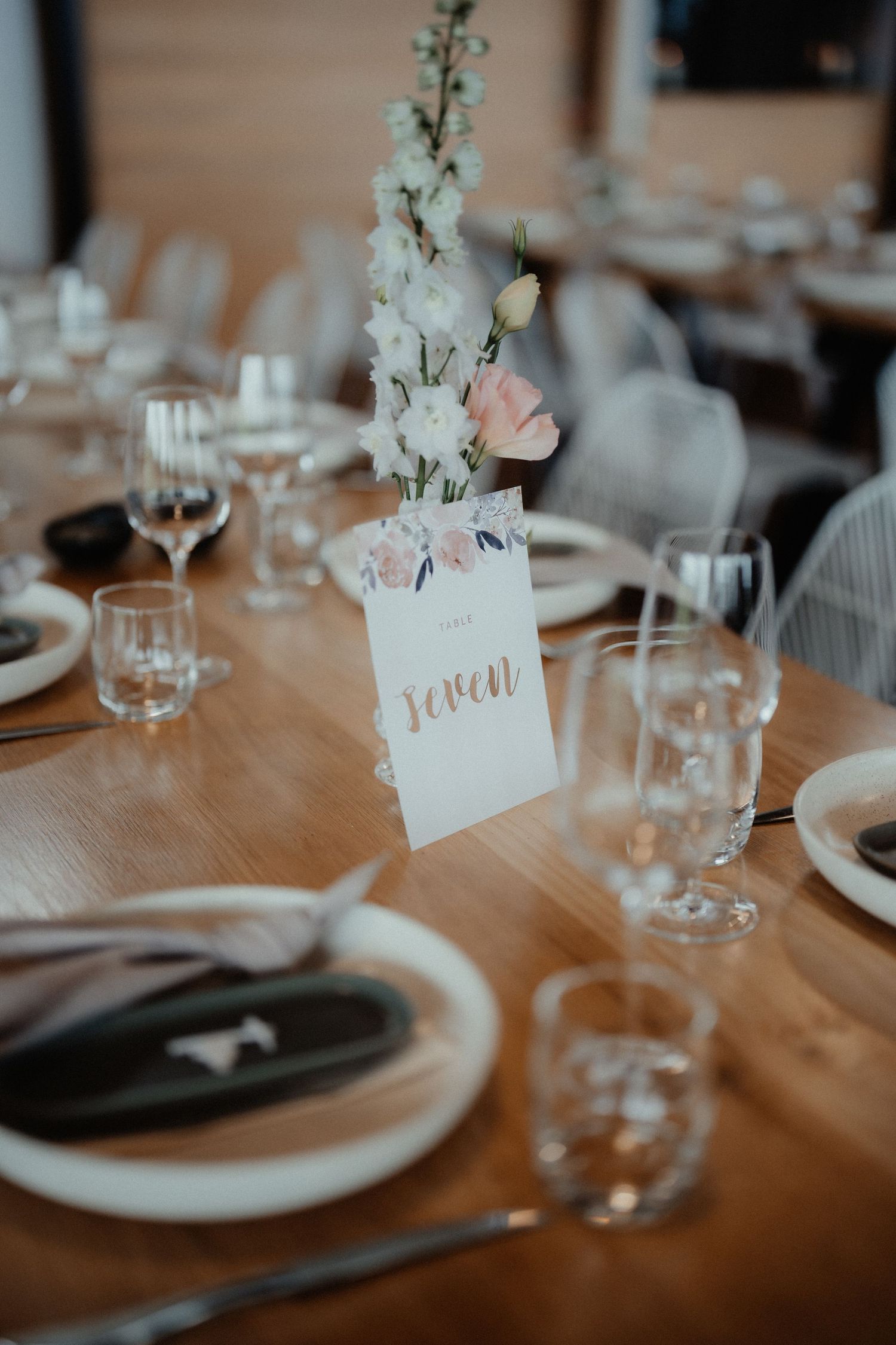 Wedding reception table with a white floral arrangement, a handwritten table number card, wine glasses and place settings.