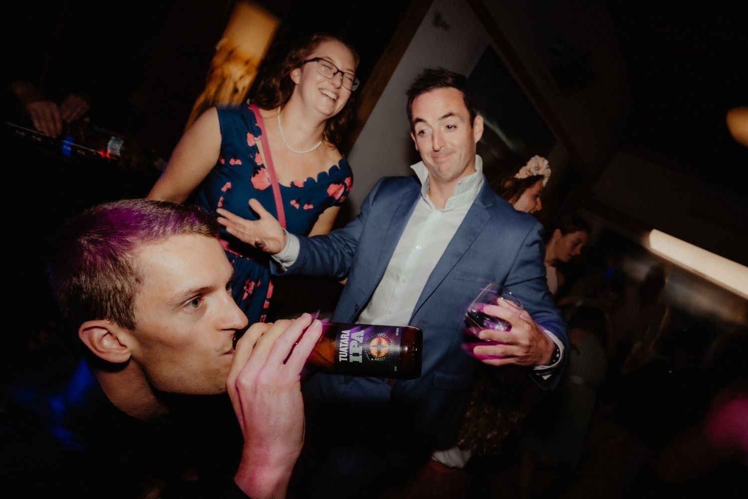 Guests celebrate at a wedding party as a man drinks from a bottle while a smiling woman and man dance nearby.