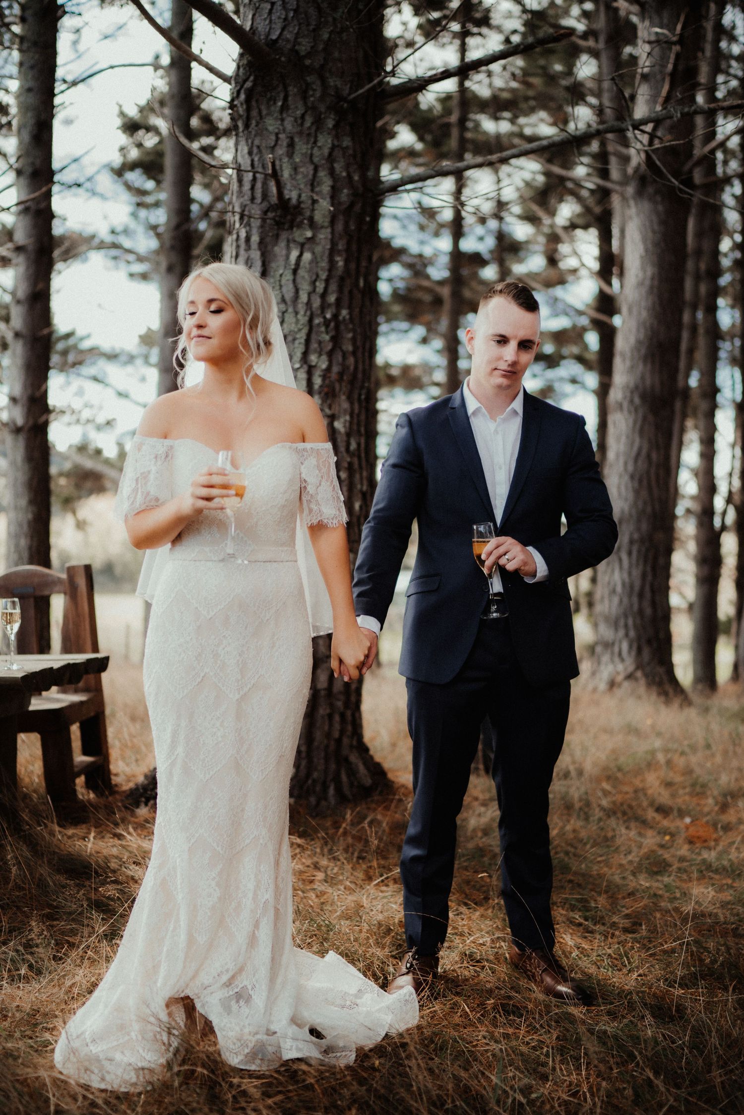 Bride in a lace gown and groom in a navy suit hold hands and champagne glasses beside a tall pine tree.
