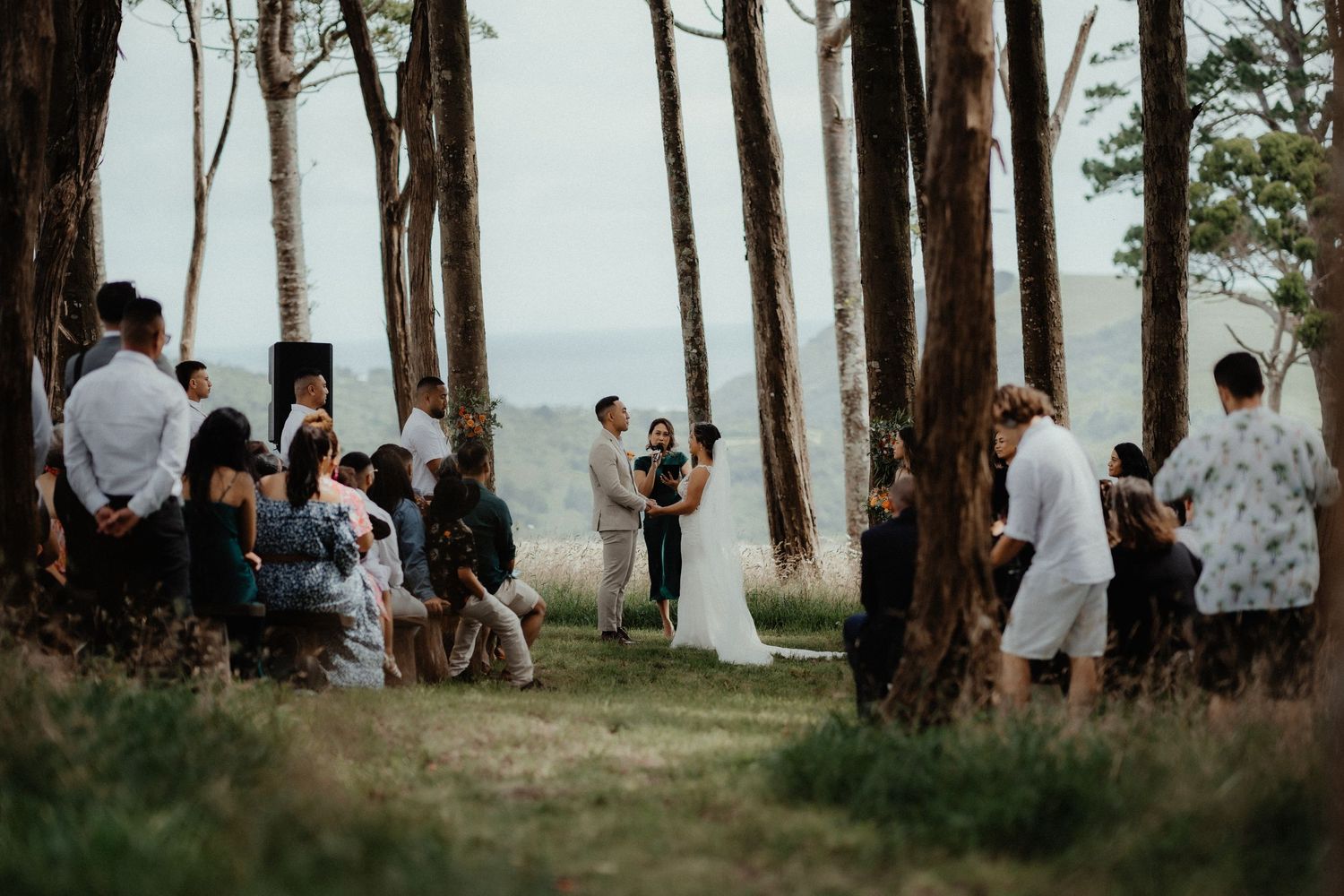 Wedding guests sit under tall pine trees watching a bride and groom exchange vows at an outdoor ceremony with a distant ocean view.