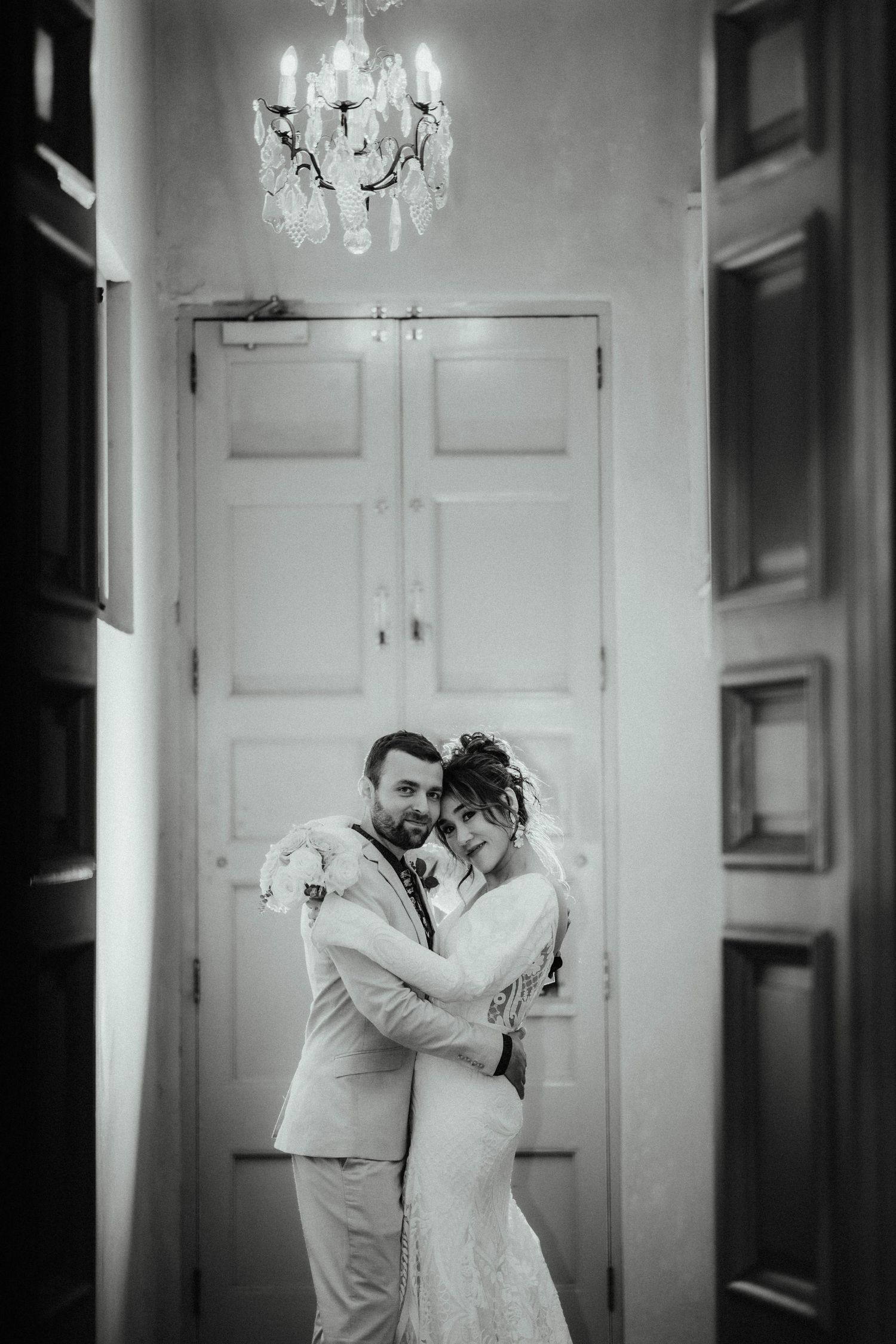Black and white portrait of a happy bride and groom embracing in a hallway beneath a vintage chandelier.