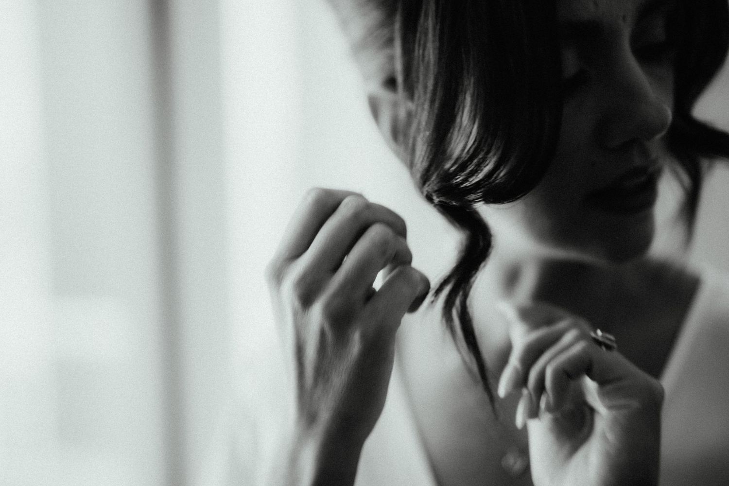 Black and white close-up of a bride's hands adjusting her hair, with a ring visible on her finger near a bright window.