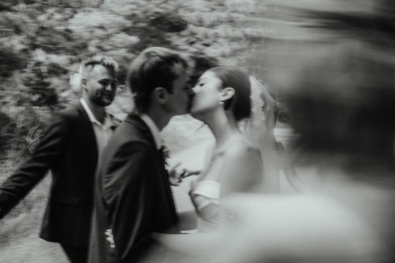 Black and white motion-blurred photo of a bride and groom kissing as a smiling guest looks on during a wedding celebration.