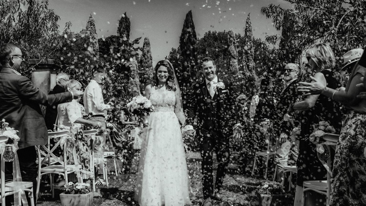 Bride in a white lace gown and groom in a dark suit walk down an outdoor aisle as guests toss confetti at their garden wedding ceremony.