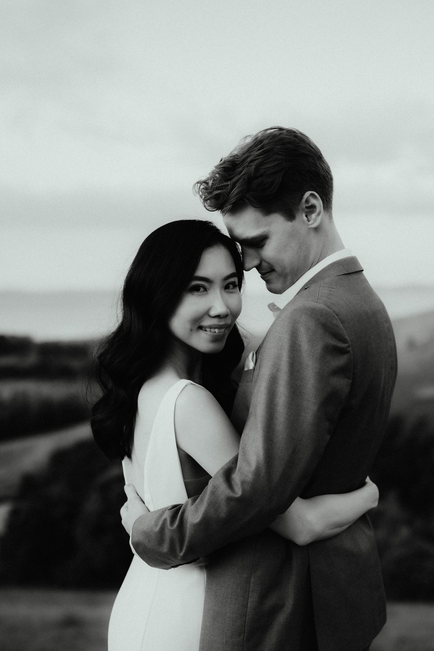 Smiling bride embraced by her groom in a black and white engagement portrait with rolling hills in the background.