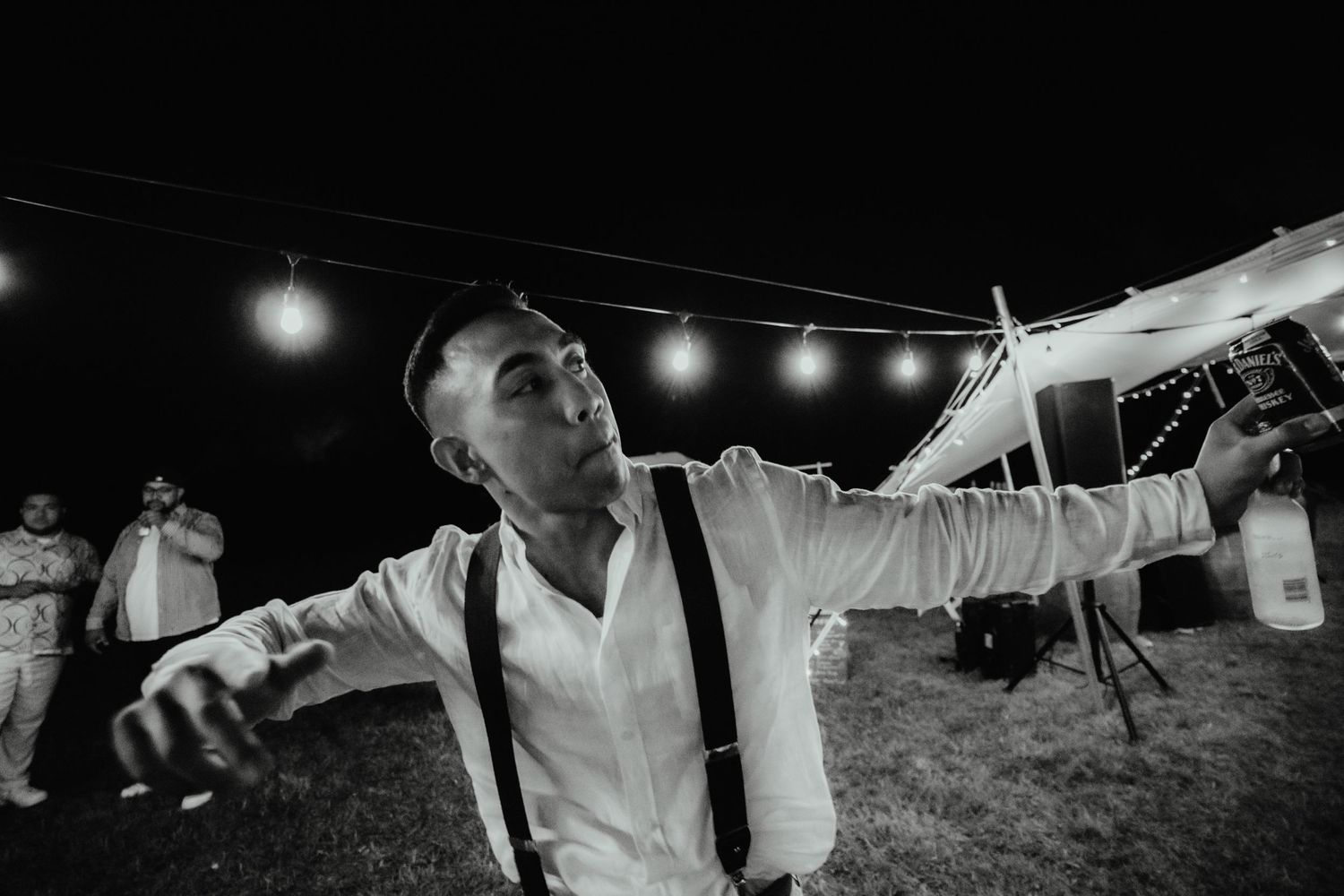 Groom in white shirt and suspenders gestures playfully at an evening outdoor wedding reception beneath string lights and a marquee tent.