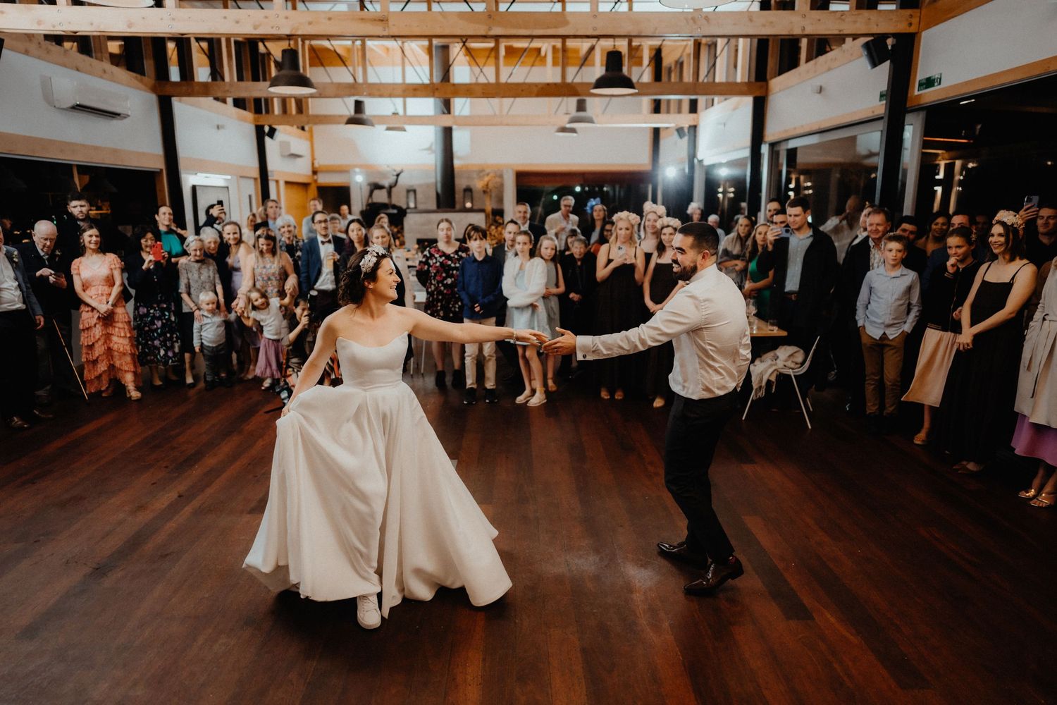 Bride and groom share their first dance on a wooden floor while wedding guests watch and gather around them.