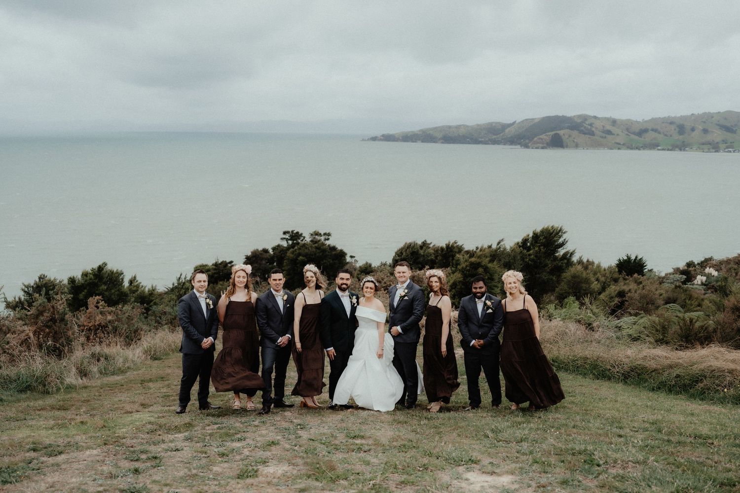 Bride, groom and wedding party in dark autumn tones pose together on a grassy coastal hilltop beneath a moody overcast sky.