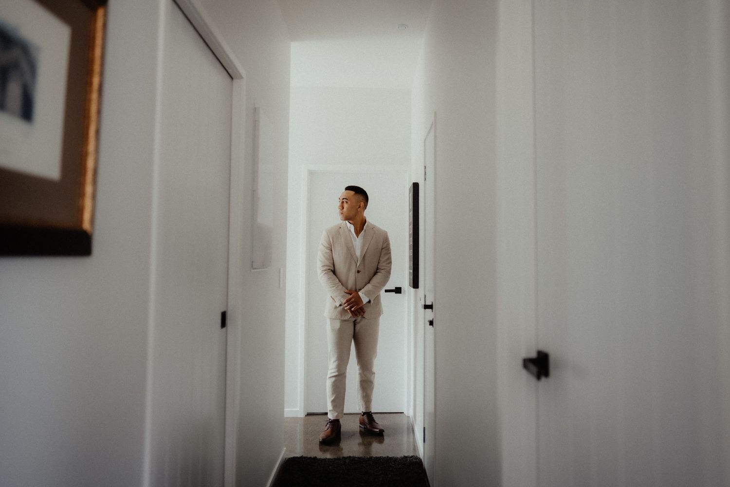A groom in a light suit stands with hands clasped in a white hallway, glancing aside before his wedding ceremony.
