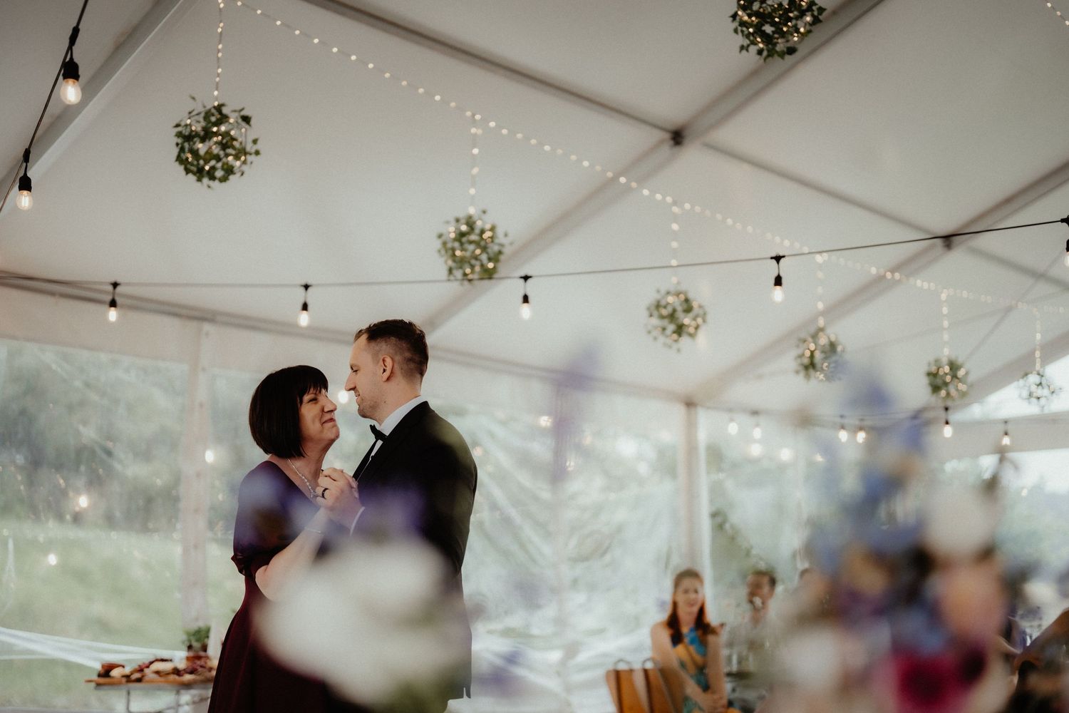 A couple shares a slow dance in a tent reception with string lights and hanging foliage balls as guests watch.
