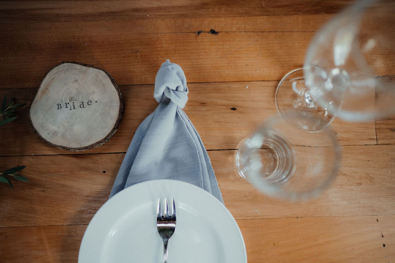 Overhead view of a rustic wedding table setting with a gray napkin, white plate, fork, wine glasses and a wooden log slice place card.