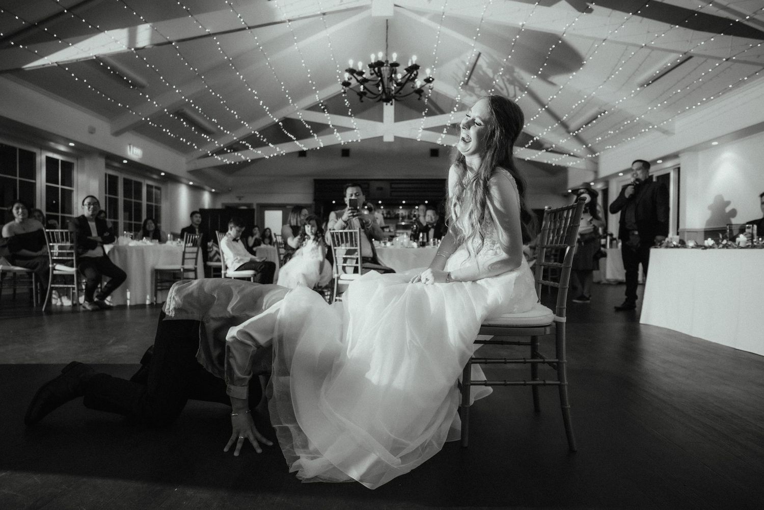 Black and white photo of a groom kneeling to remove the bride's garter while she sits on a chair at the wedding reception.
