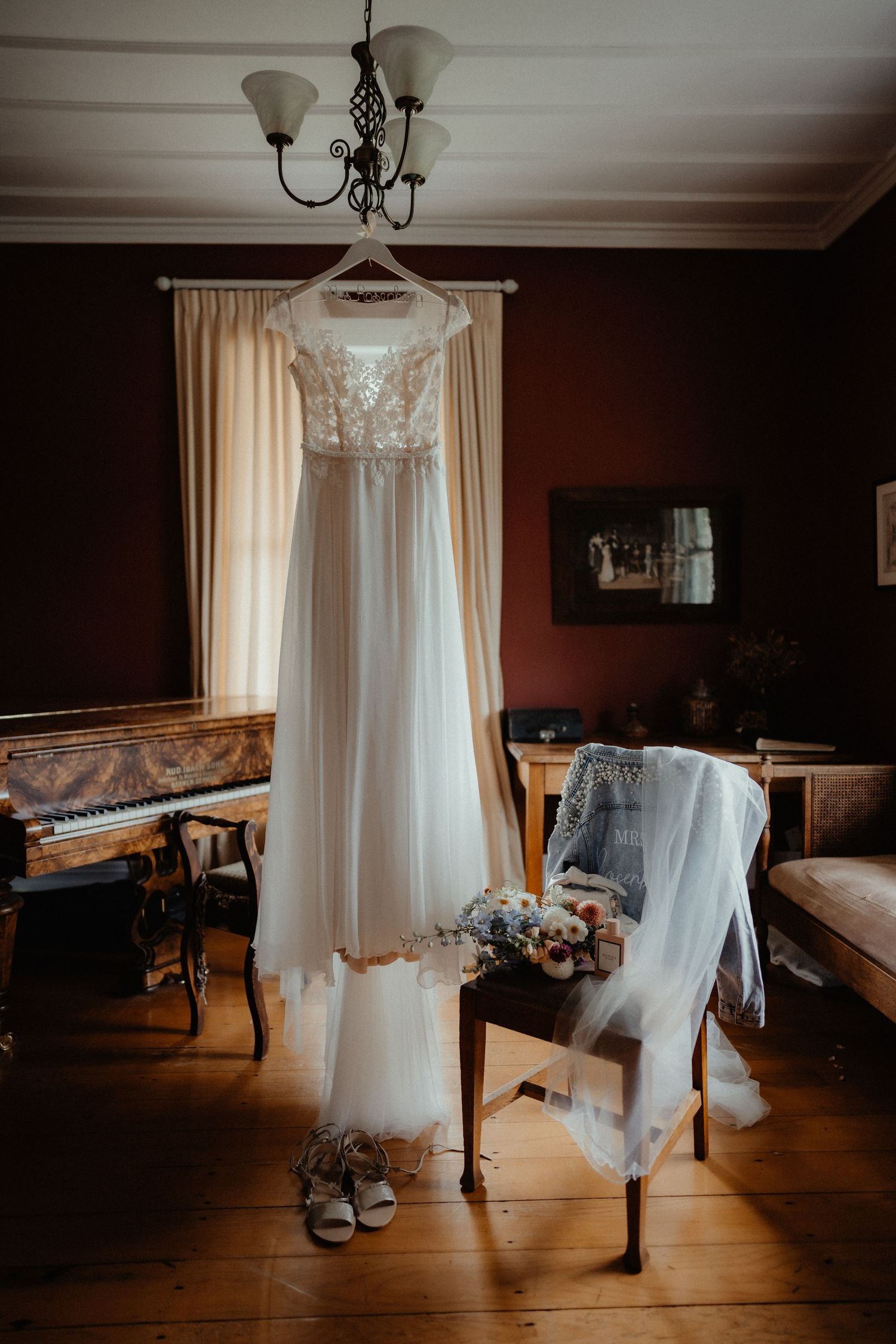 Lace wedding dress hanging from a chandelier in a rustic room with a piano, bridal bouquet, veil and shoes nearby.