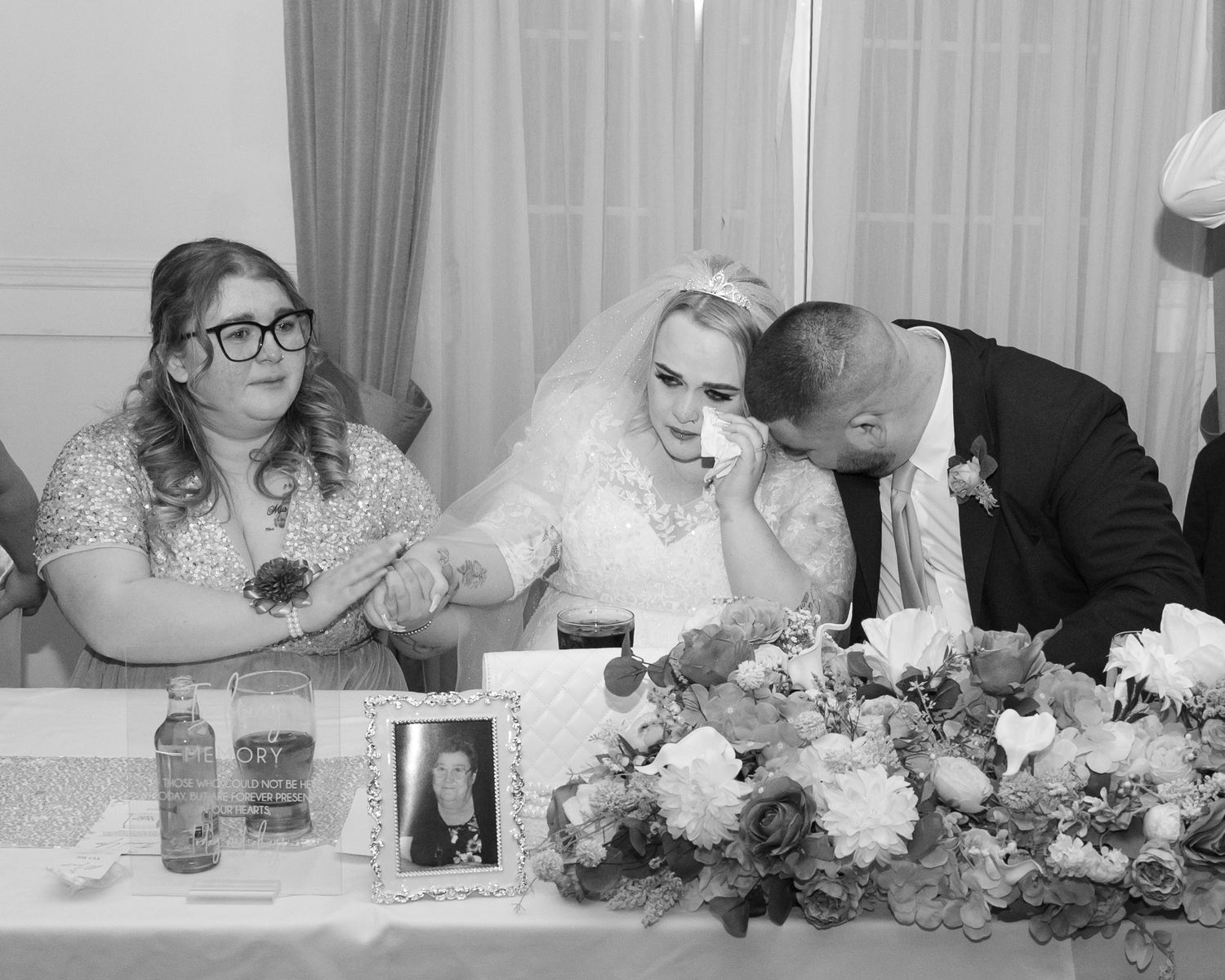 Black and white wedding photo of emotional bride and groom signing register with bridesmaid, floral arrangements on table.