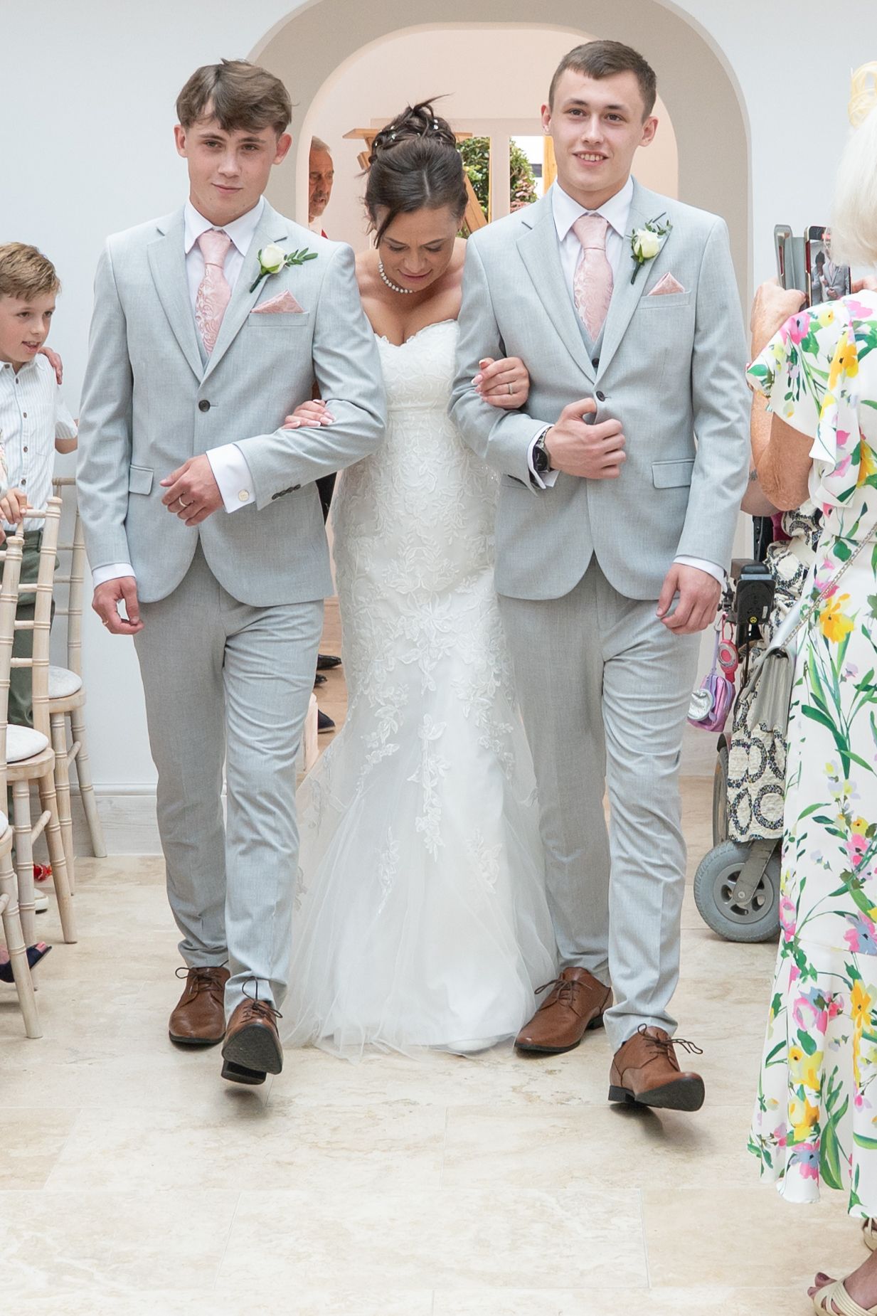 Wedding party members in light gray suits and a white dress walk down a floral-lined aisle.