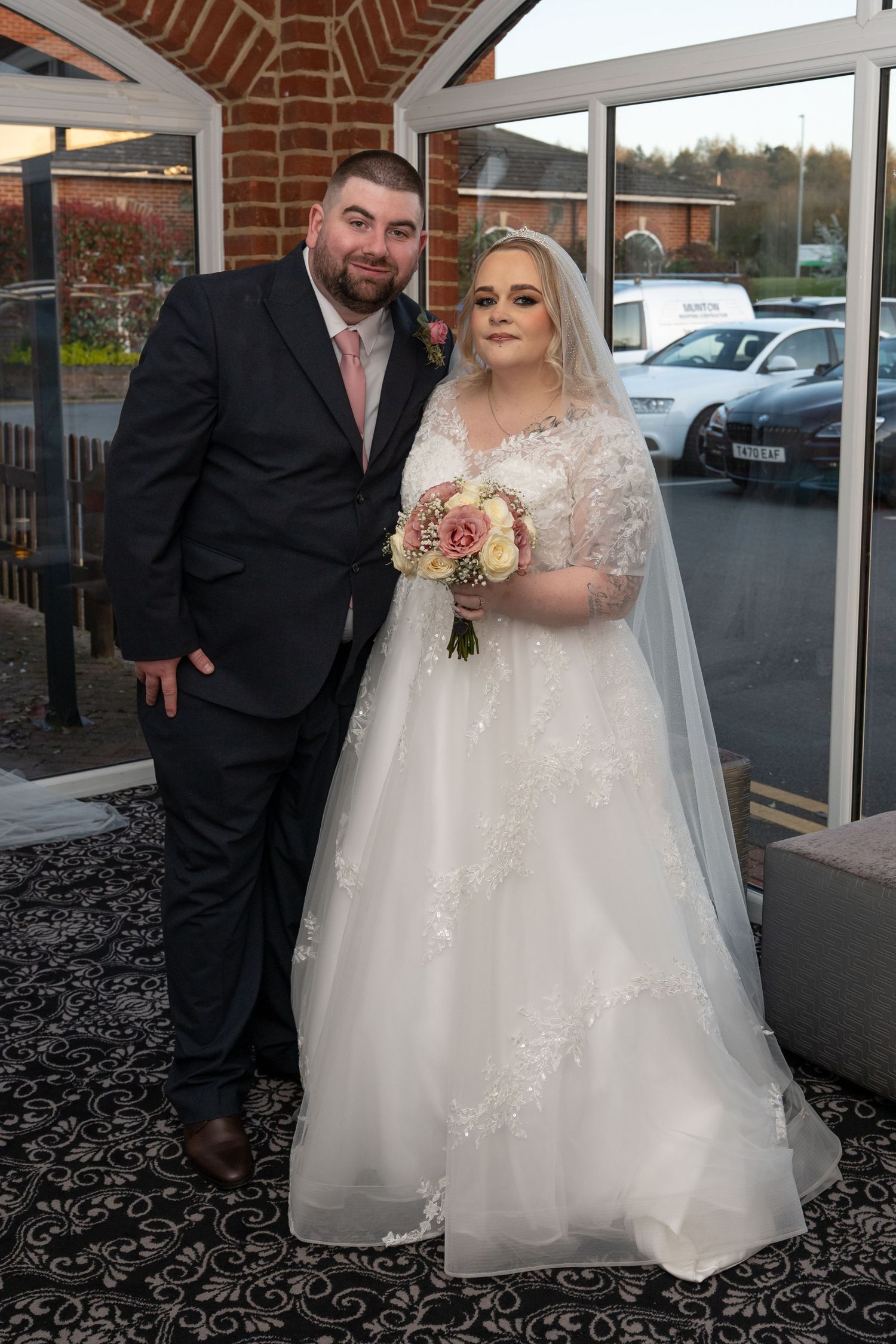 Bride in white lace gown holding pink bouquet stands with groom in dark suit outside wedding venue.