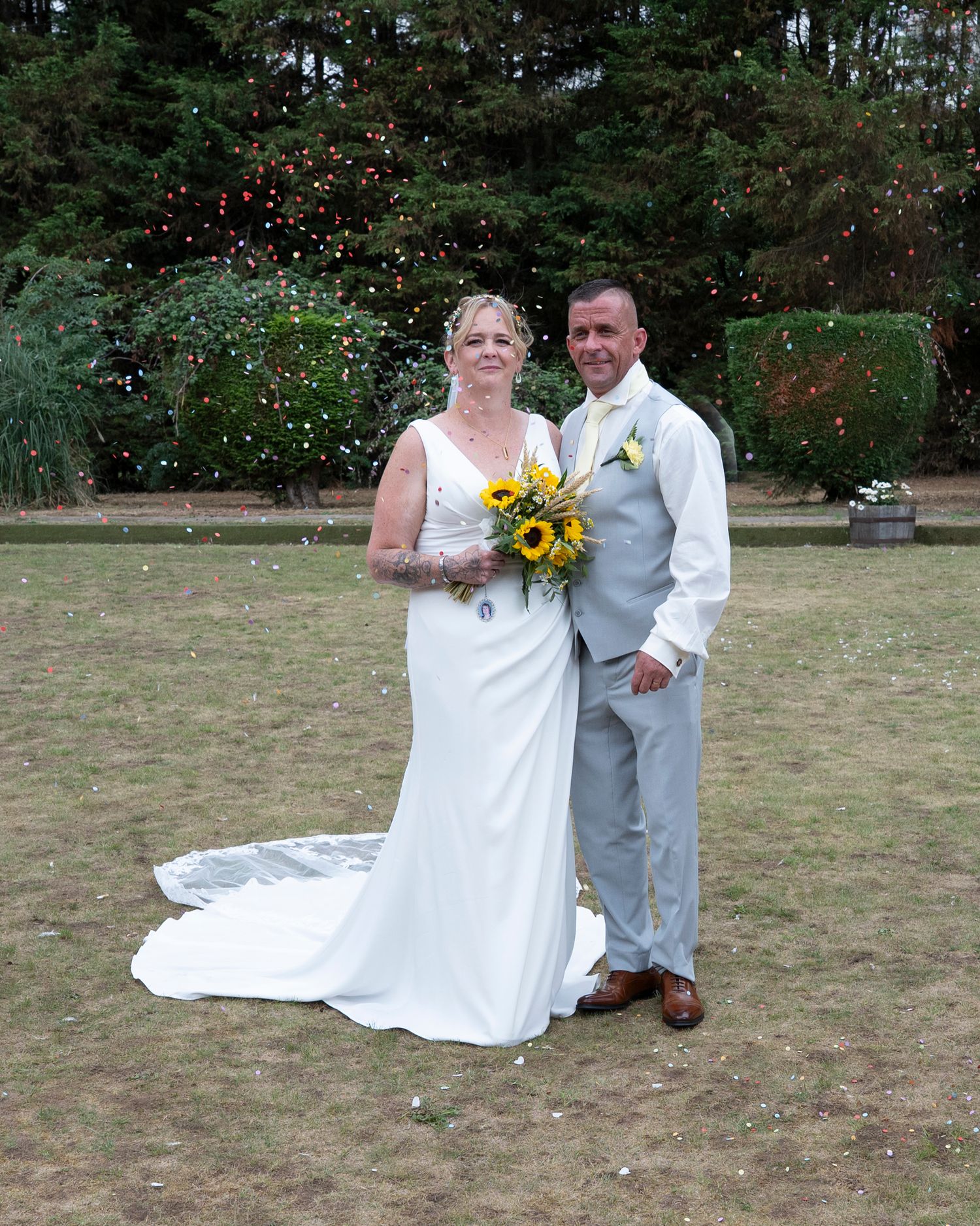 Wedding couple poses with sunflower bouquet in an outdoor garden setting.
