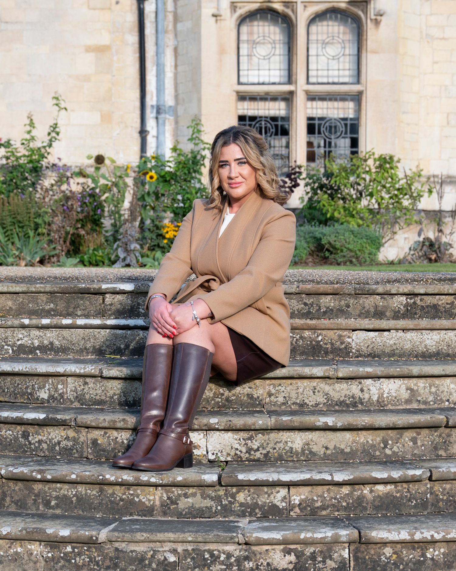Person in brown coat and boots sits on stone steps outside historic building.