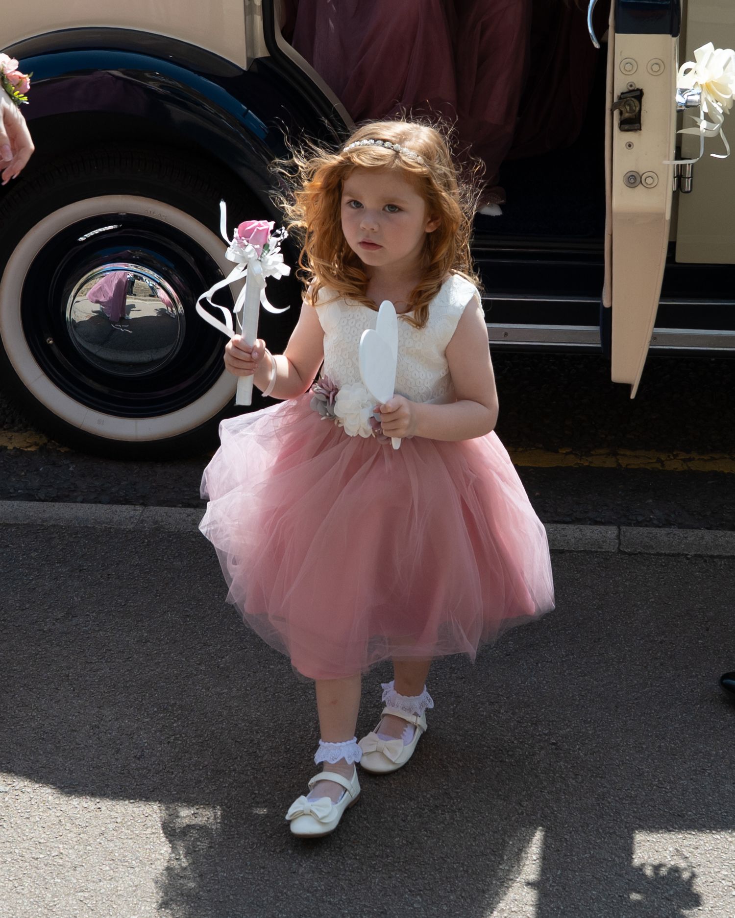 A young flower girl in a pink tulle dress and white shoes crouches down.