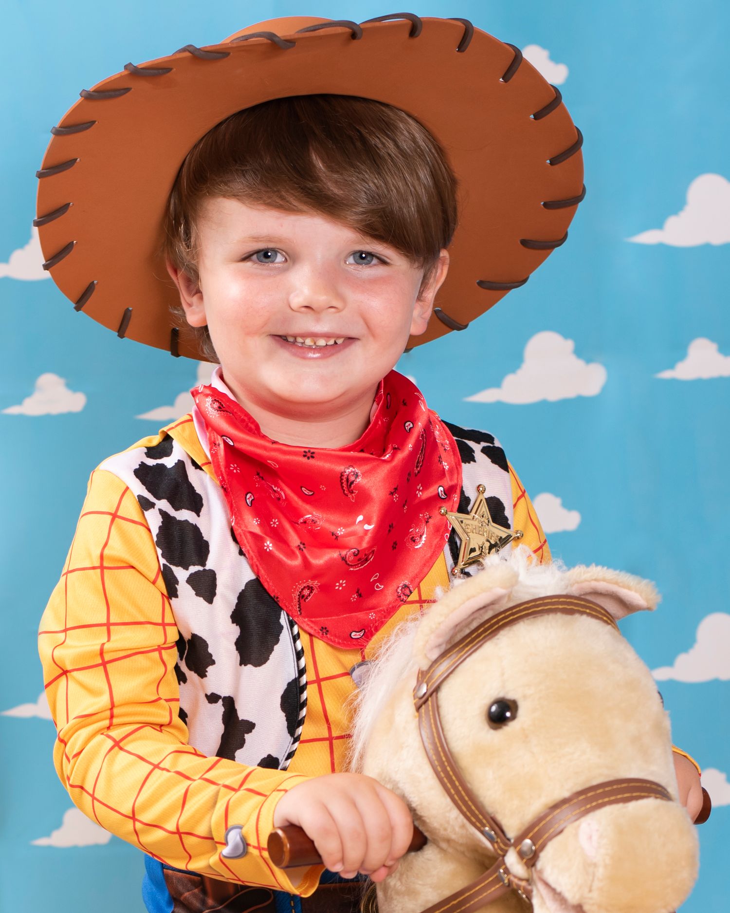 A child dressed in a Woody from Toy Story costume with cowboy hat and red bandana poses with a toy horse.