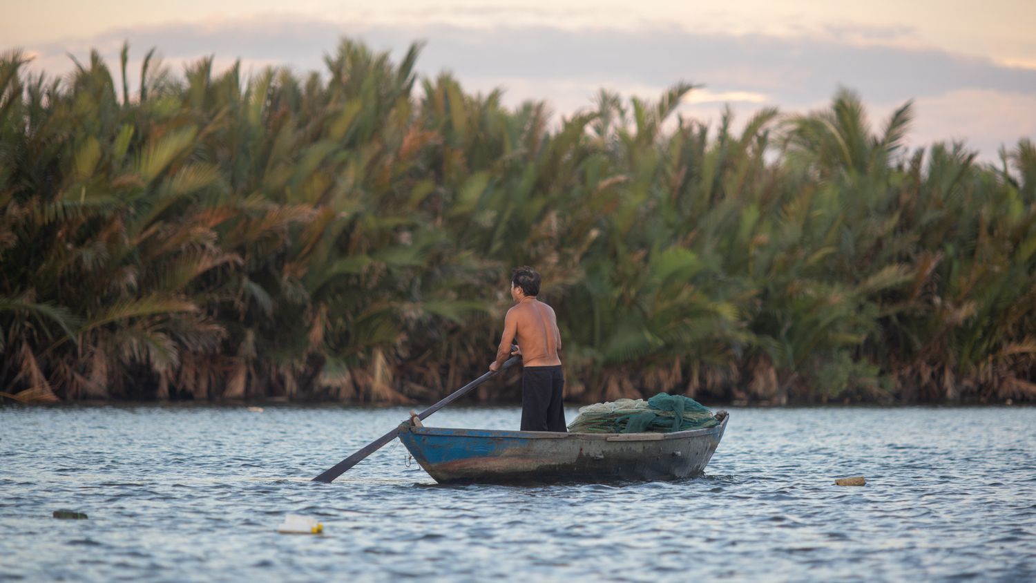 Shirtless man rowing a small wooden boat on a calm river surrounded by palm trees.