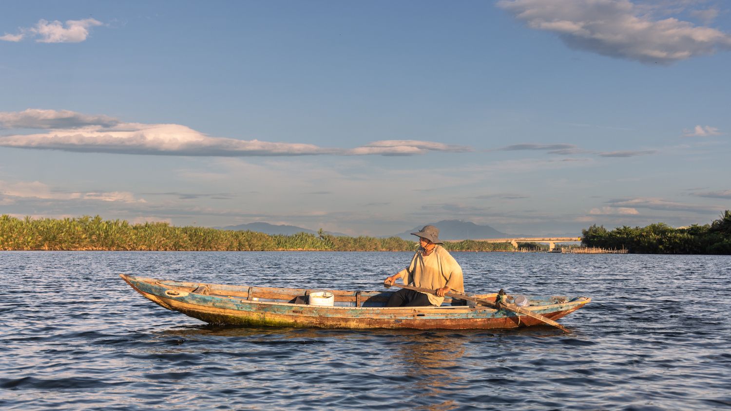 Elderly fisherman sitting in a traditional wooden boat on a wide calm river.