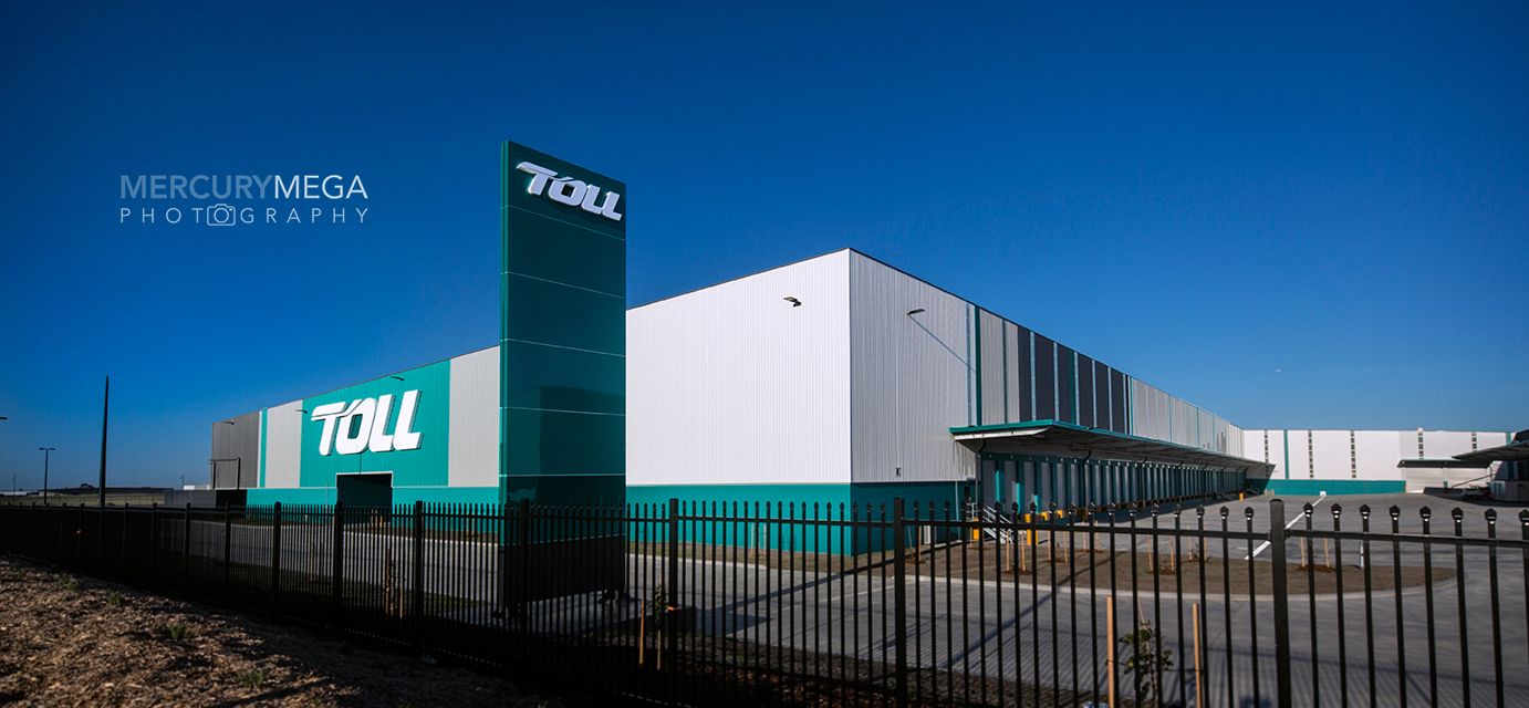 Toll logistics warehouse exterior featuring teal and white building facade with prominent Toll signage under clear blue sky.
