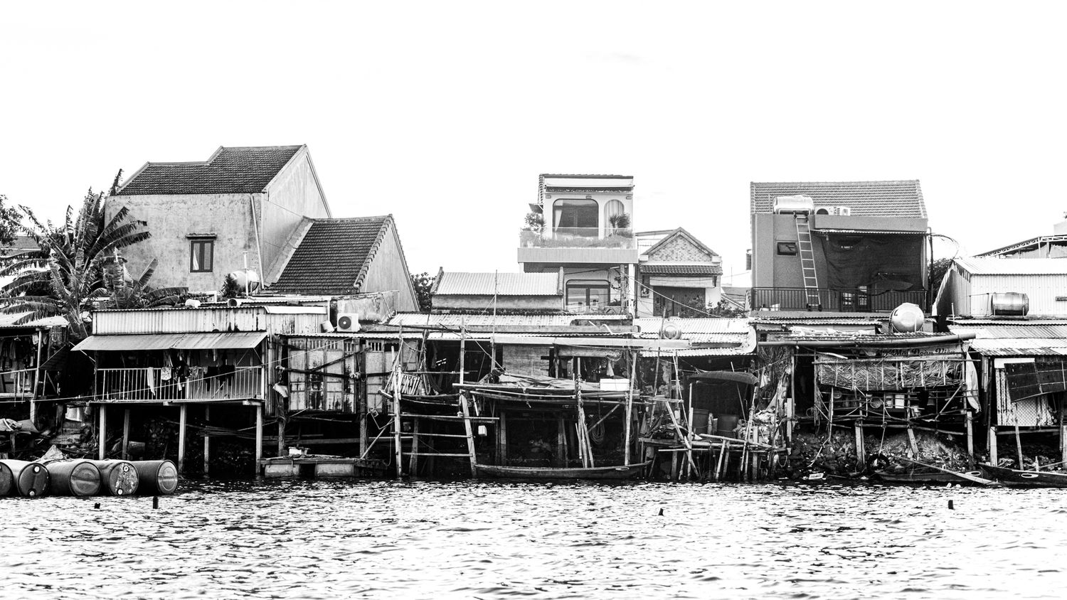 Black and white photo of waterfront stilt buildings along a busy riverside town.