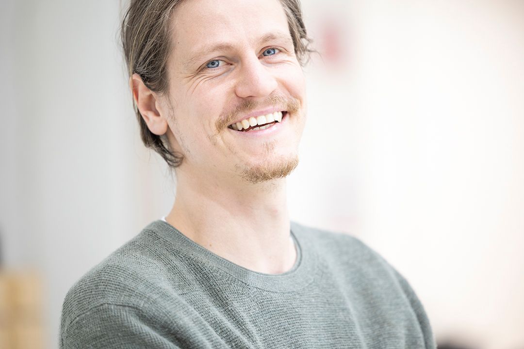 Smiling young man with brown hair and goatee wearing a gray sweater in a bright indoor setting.