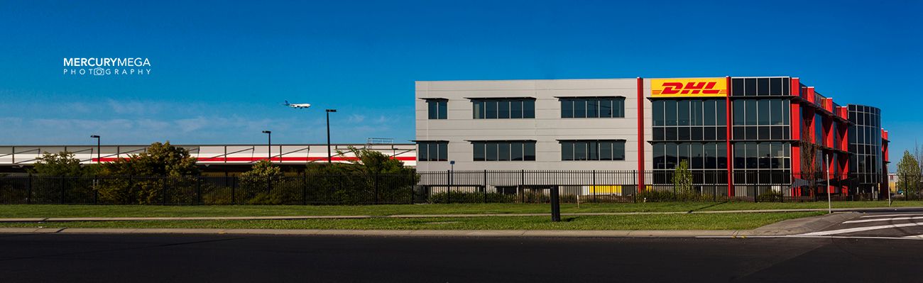Panoramic view of a modern DHL logistics facility with red and yellow branding, featuring a sleek office building and warehouse.