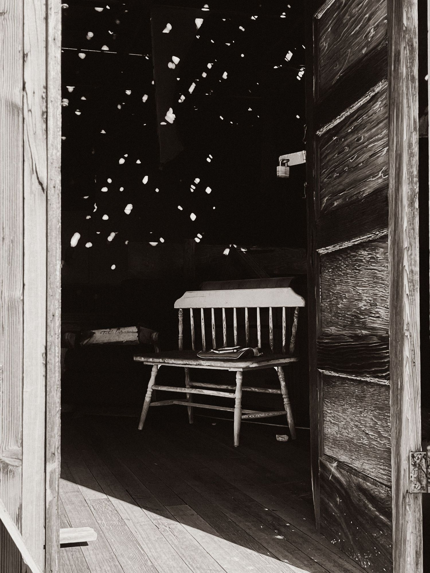 Black and white documentary photograph of an empty wooden chair inside a rustic building shot on 35mm film. Goodreau.co