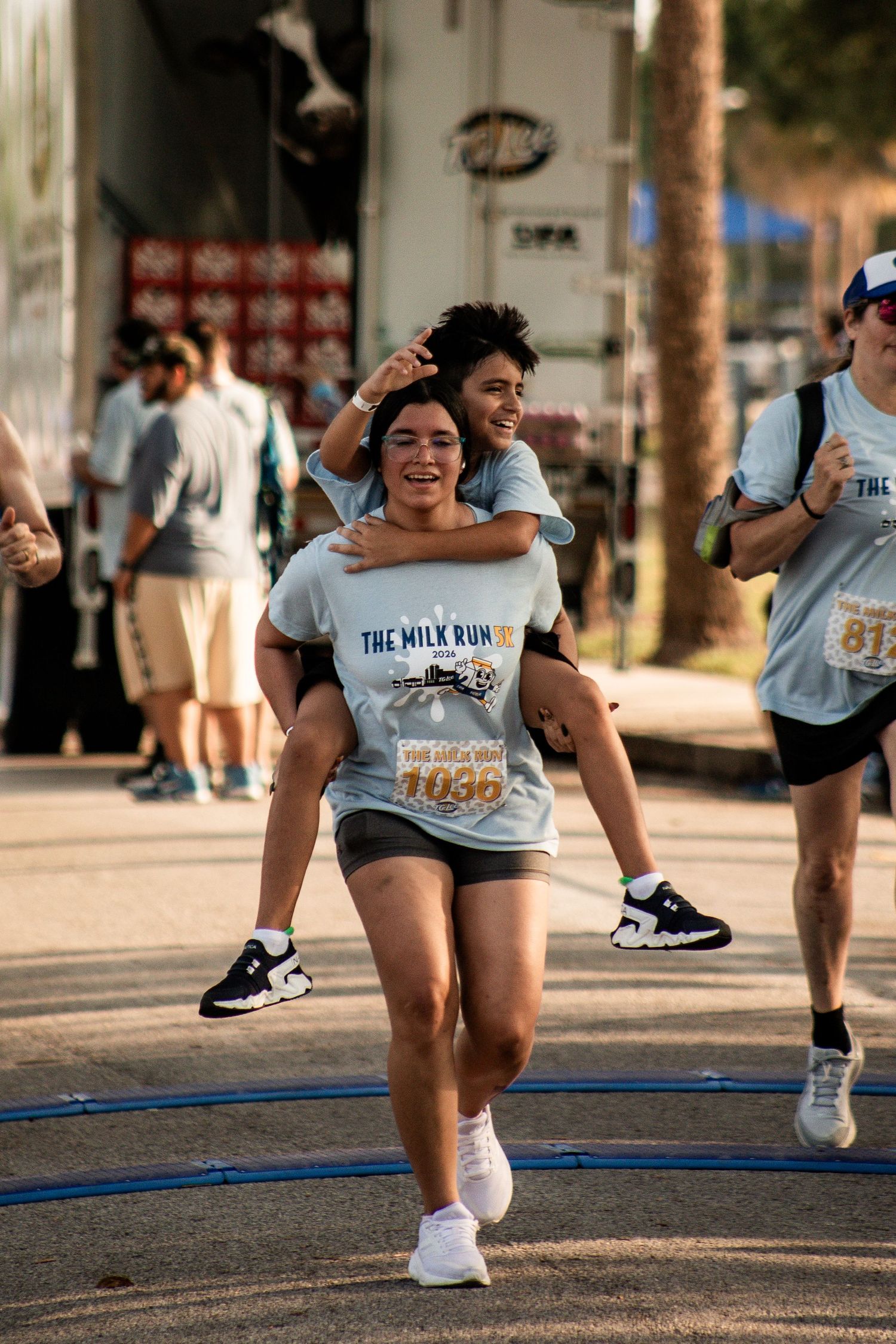 Race participant carrying another runner piggyback across the finish line at a community 5K. Goodreau.co