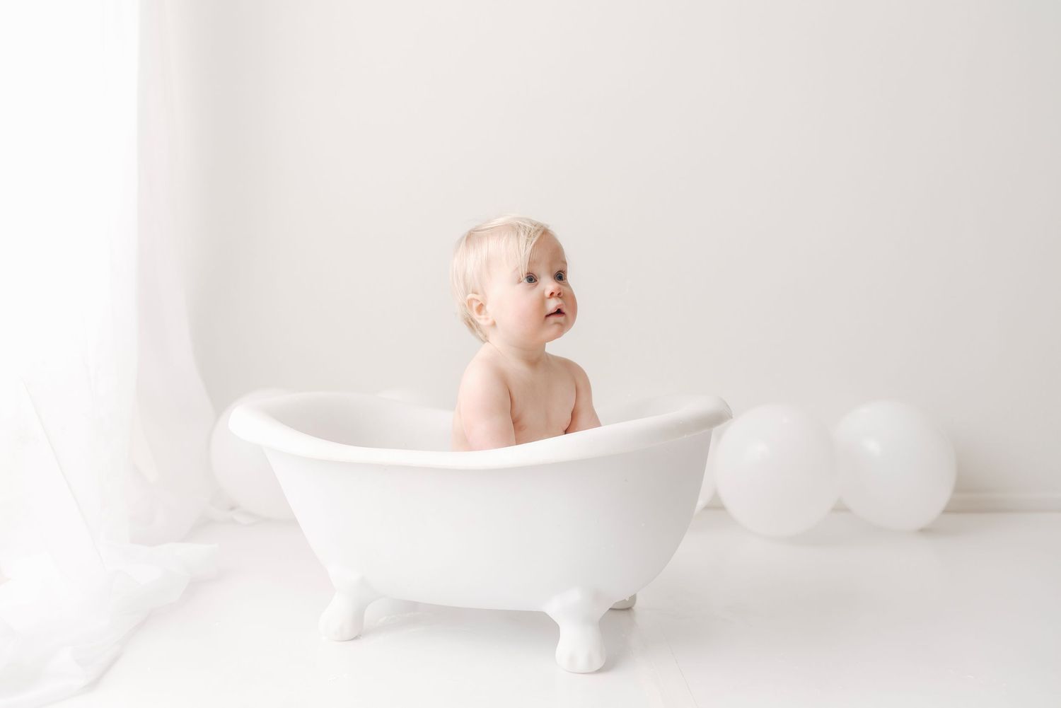 A cute baby sits in a small white clawfoot bathtub during a cake smash cleanup photo session.