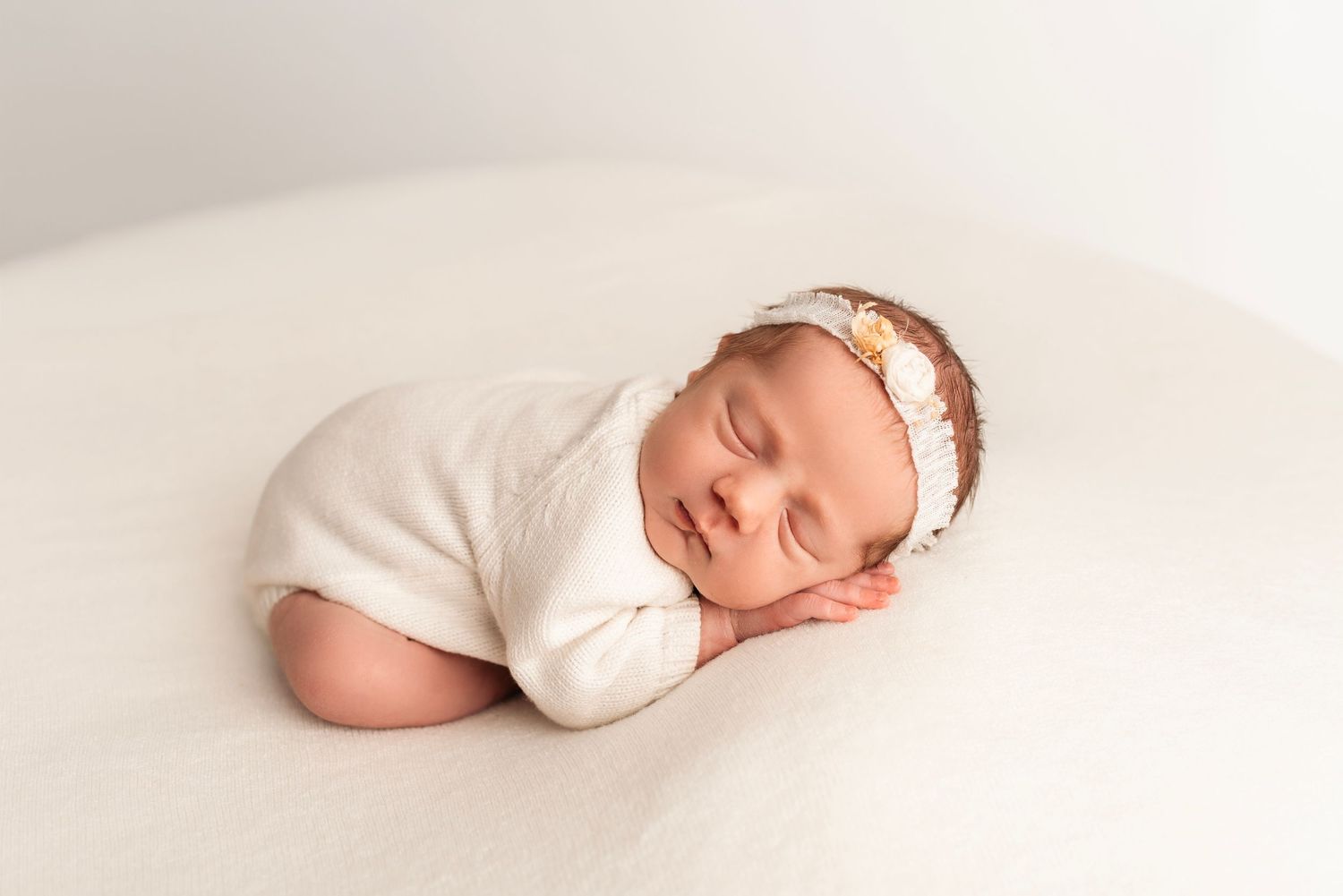 Peaceful sleeping newborn baby in white outfit and floral headband posed on soft white blanket.