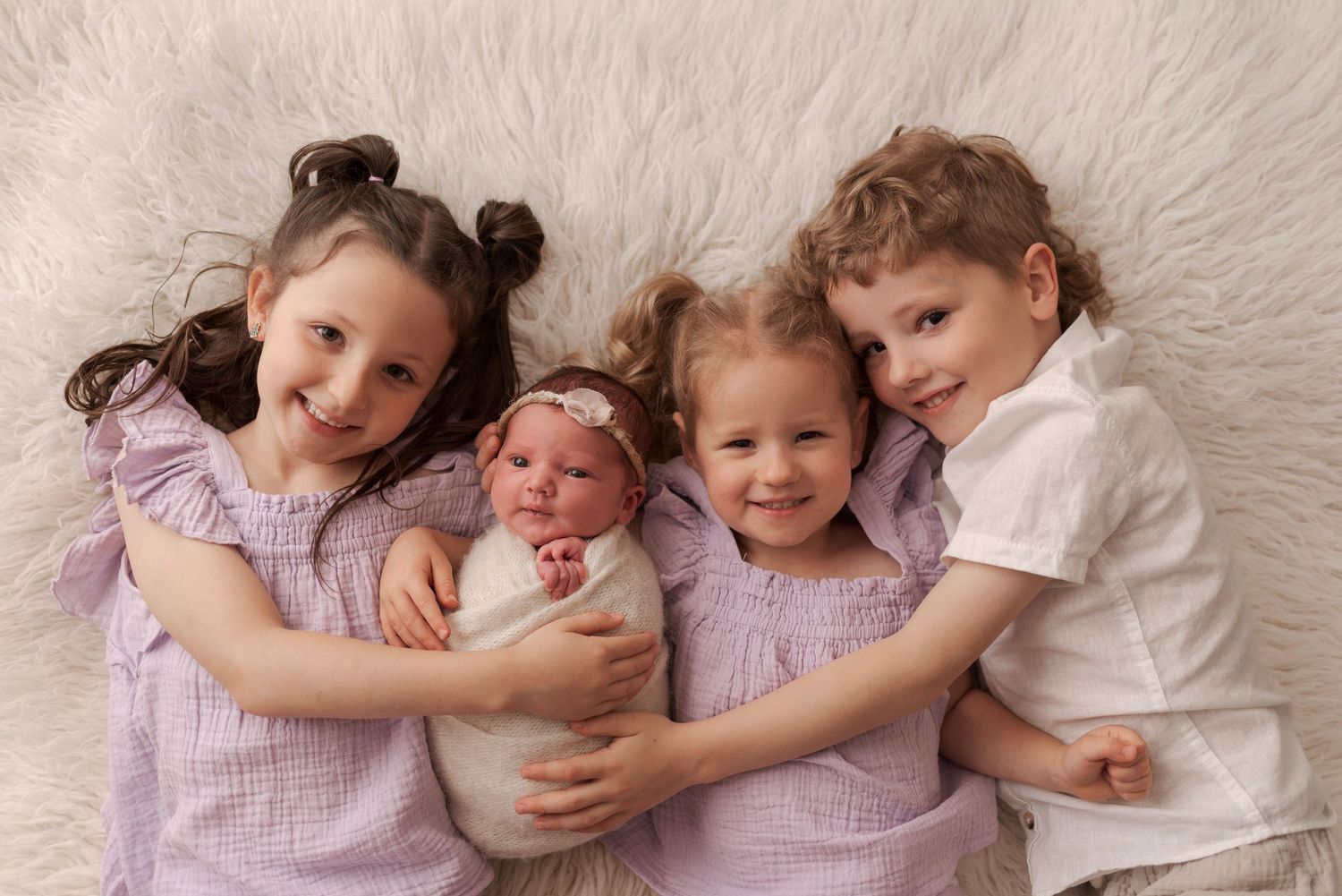 Four siblings pose together on a fluffy white rug, with three older children lovingly holding a swaddled newborn baby.