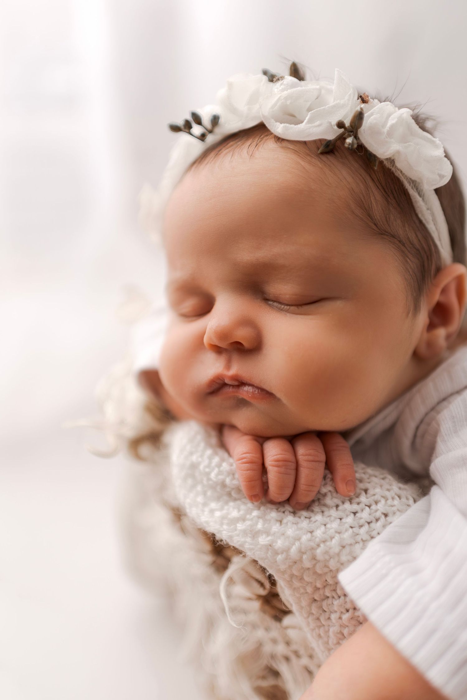 Close-up portrait of sleeping newborn baby girl wearing white floral headband on soft blanket.