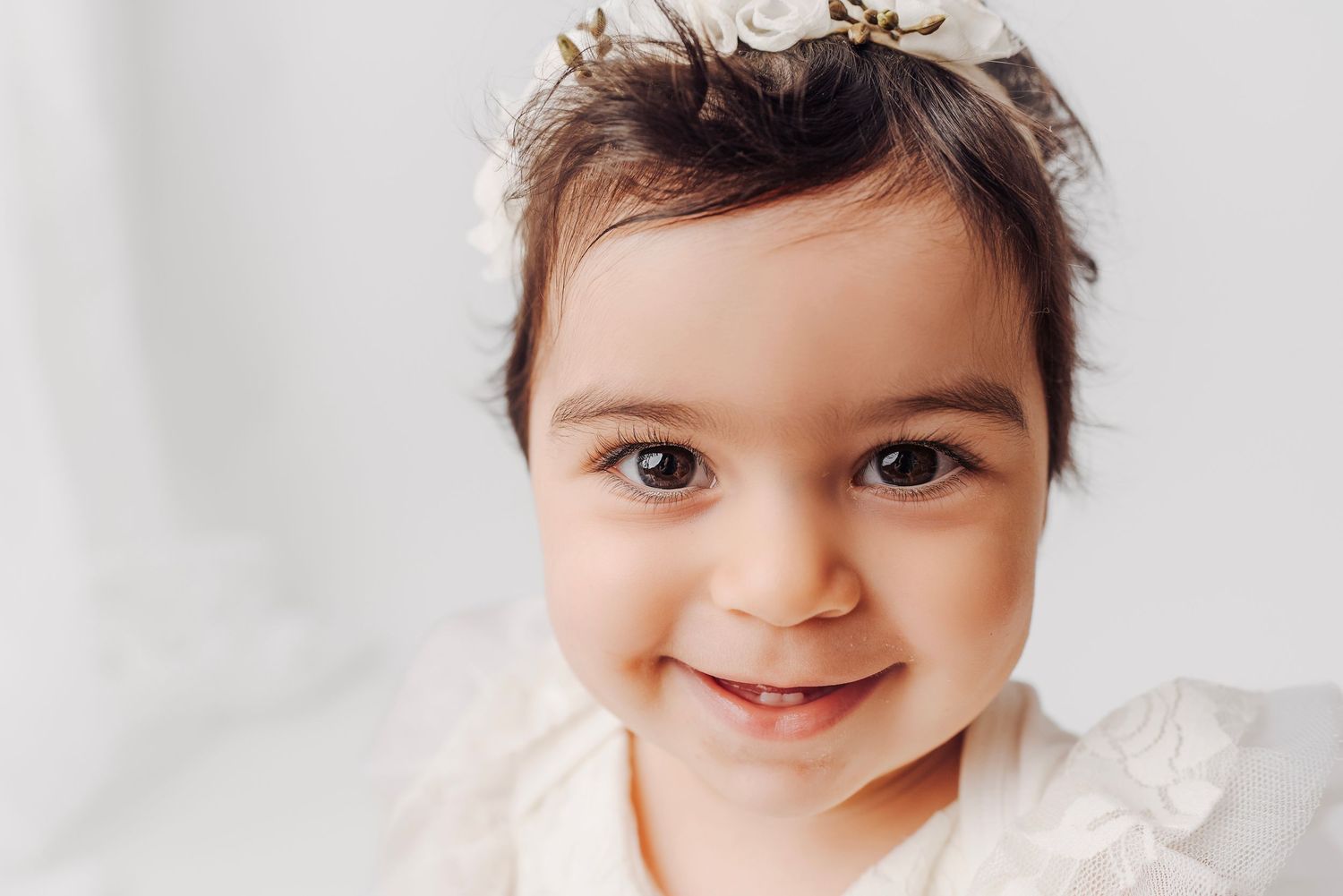 Adorable baby girl smiling with dark eyes wearing a delicate floral crown headband in a white studio.