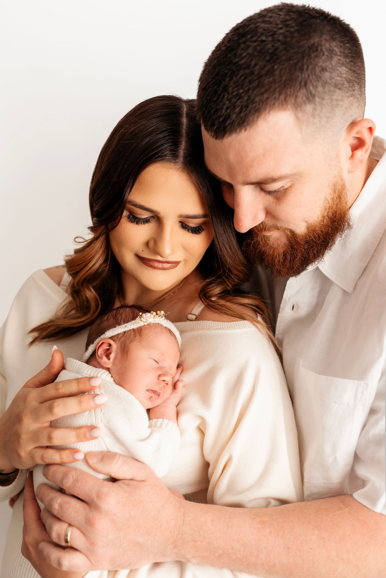 New parents in white outfits lovingly holding their sleeping newborn baby girl with floral headband.