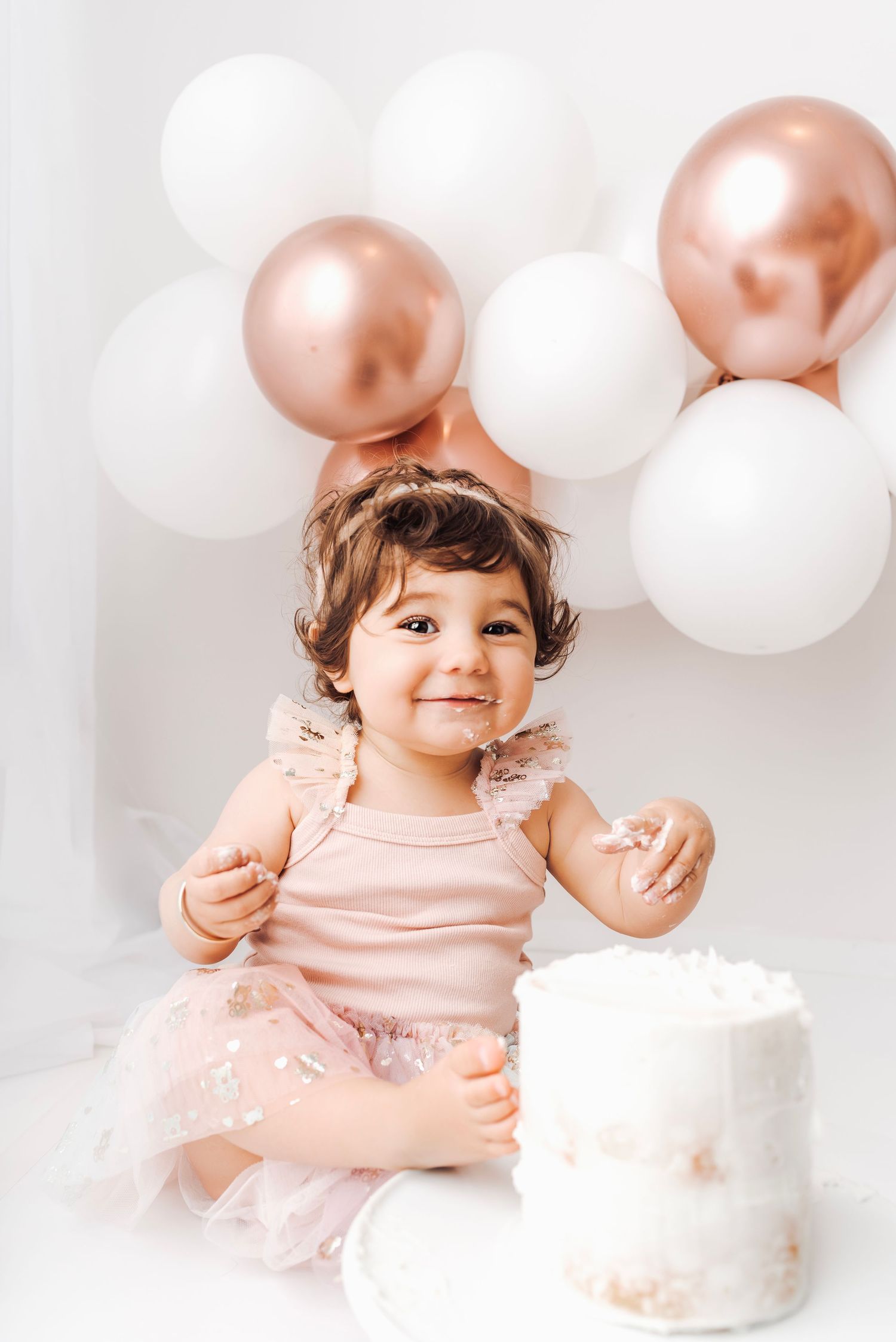 A smiling toddler girl in a pink dress sits beside a white cake with rose gold and white balloons behind her.