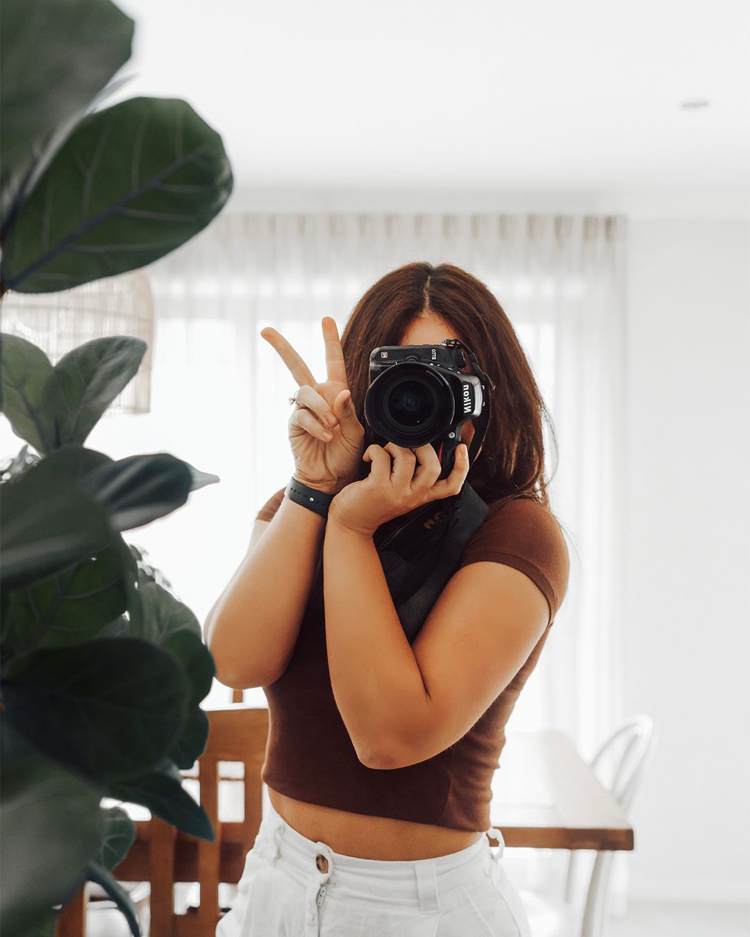 Woman holding camera up to face making peace sign in mirror selfie with indoor plant.